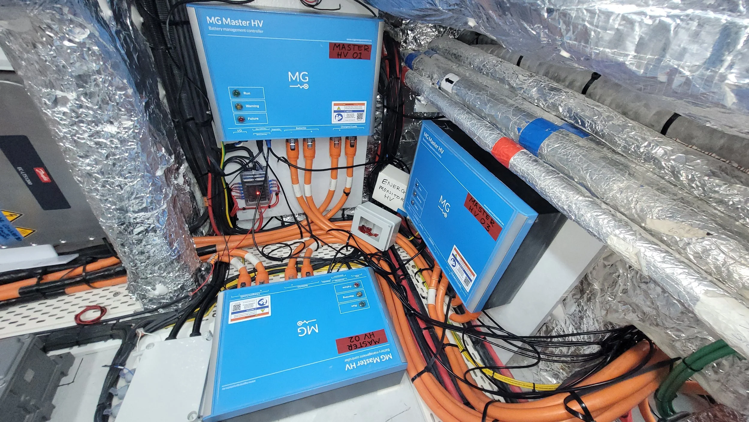 Electrical and battery management system with blue units labeled MG Master HV, orange cables, and metallic insulated pipes in a high-tech environment.