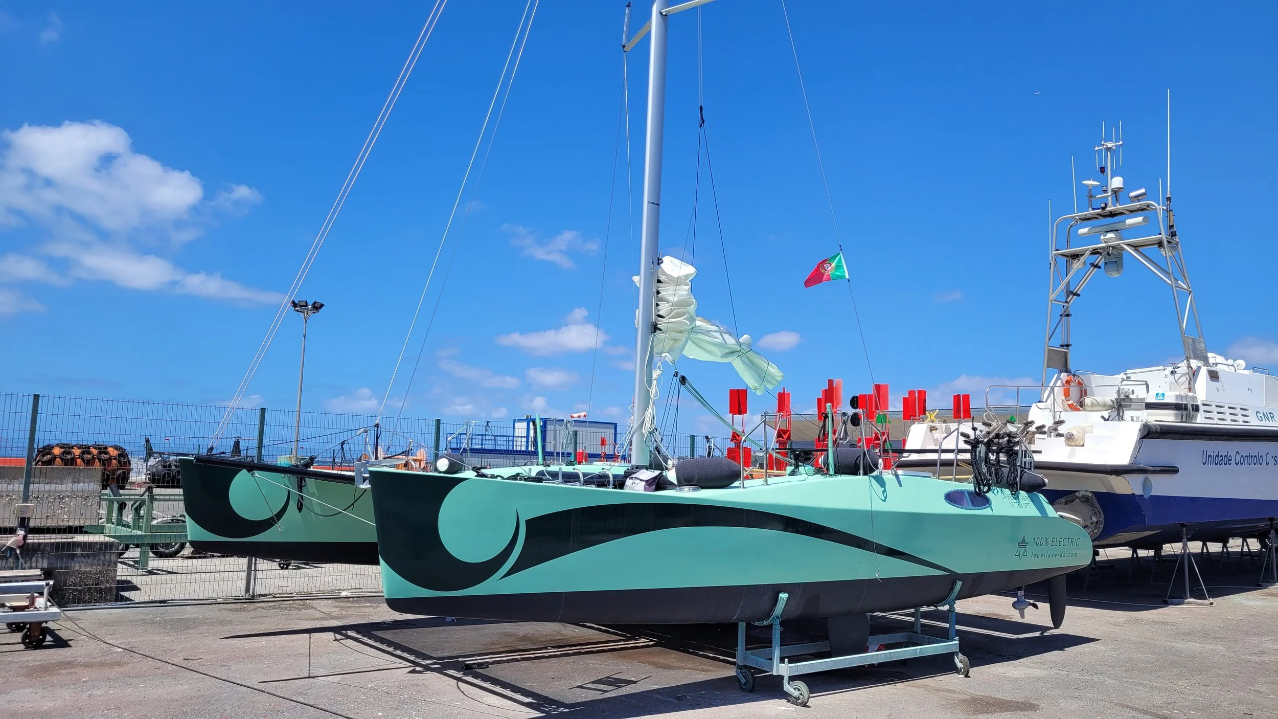 A green electric sailboat on land with a black and teal wave pattern, next to a white boat with radar and antennas, against a blue sky with clouds.