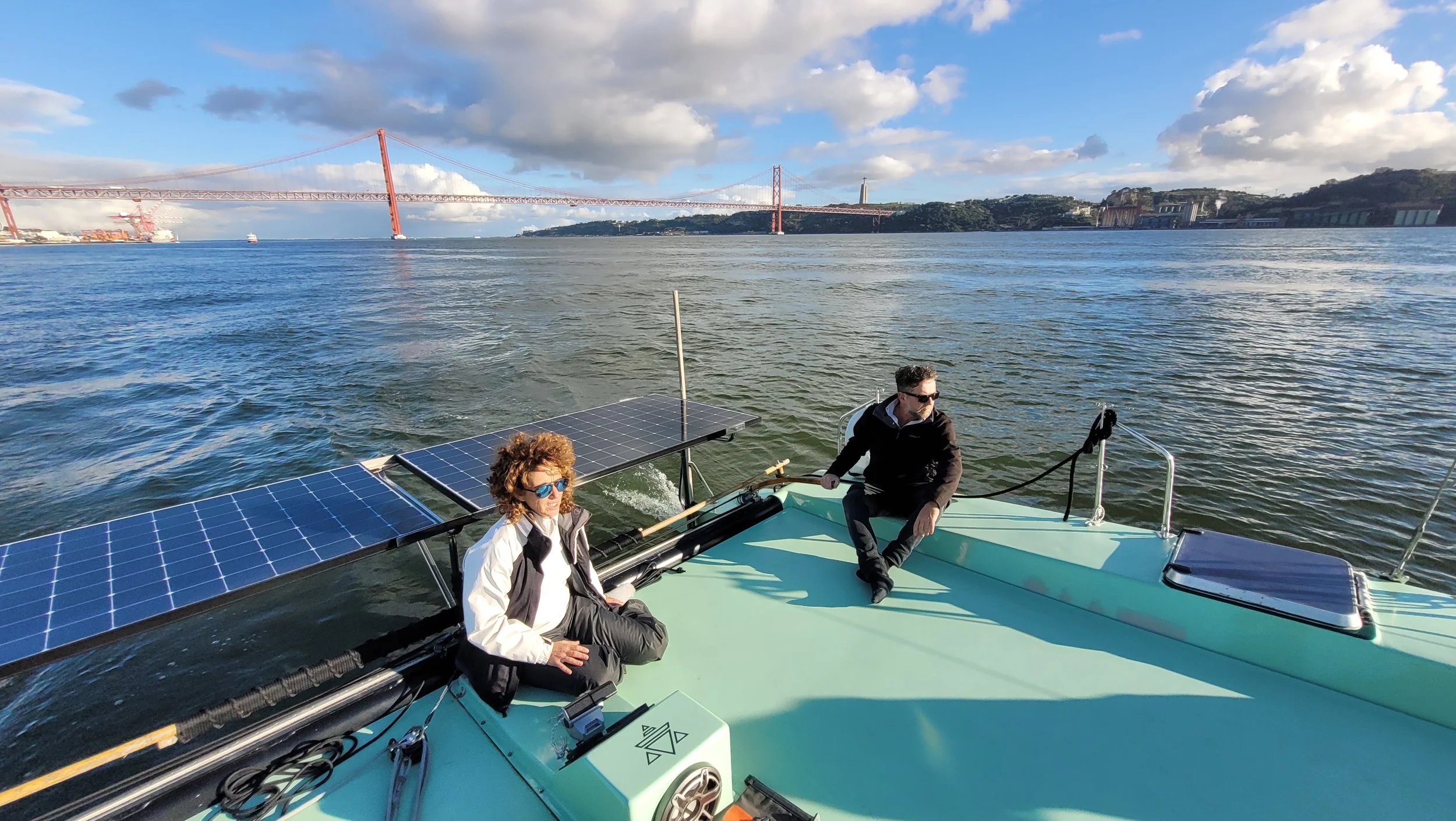 Two people sitting on the deck of a solar-powered boat with solar panels attached, on a body of water with the River Tejo in Lisbon and cityscape in the background during daytime.