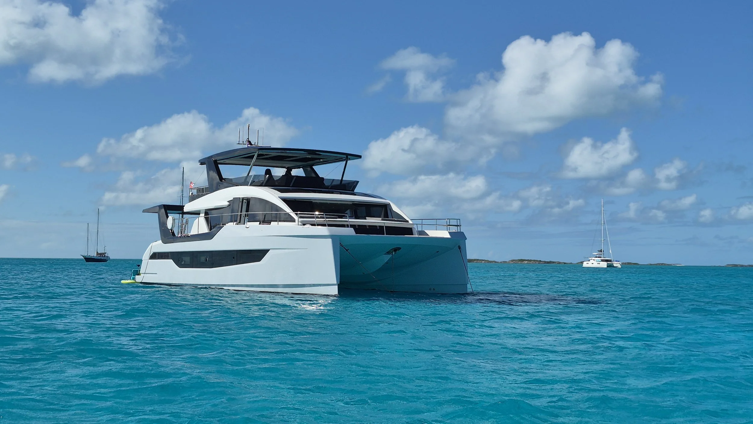 A sleek hybrid electric yacht floating on calm turquoise Bahamian water with a clear blue sky and scattered clouds in the background, and two sailboats visible in the distance.