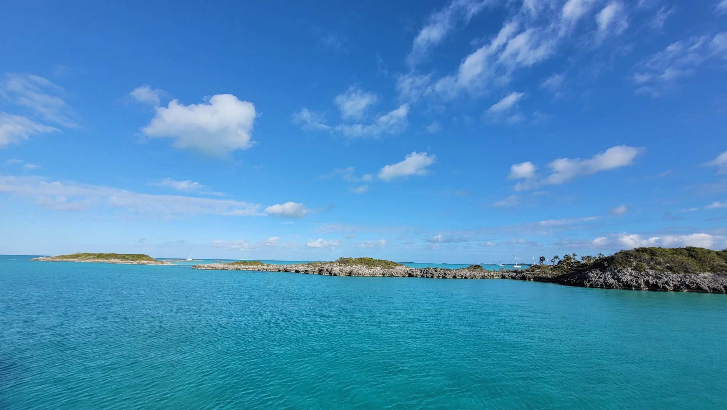 A clear blue sky with scattered clouds over calm turquoise ocean water, with small islands and rocky outcroppings in the distance.