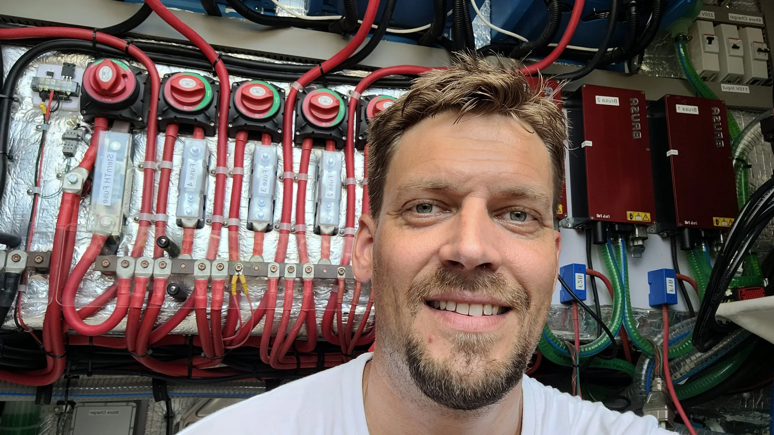 A man with short brown hair and a beard smiling in front of electrical equipment and wiring.