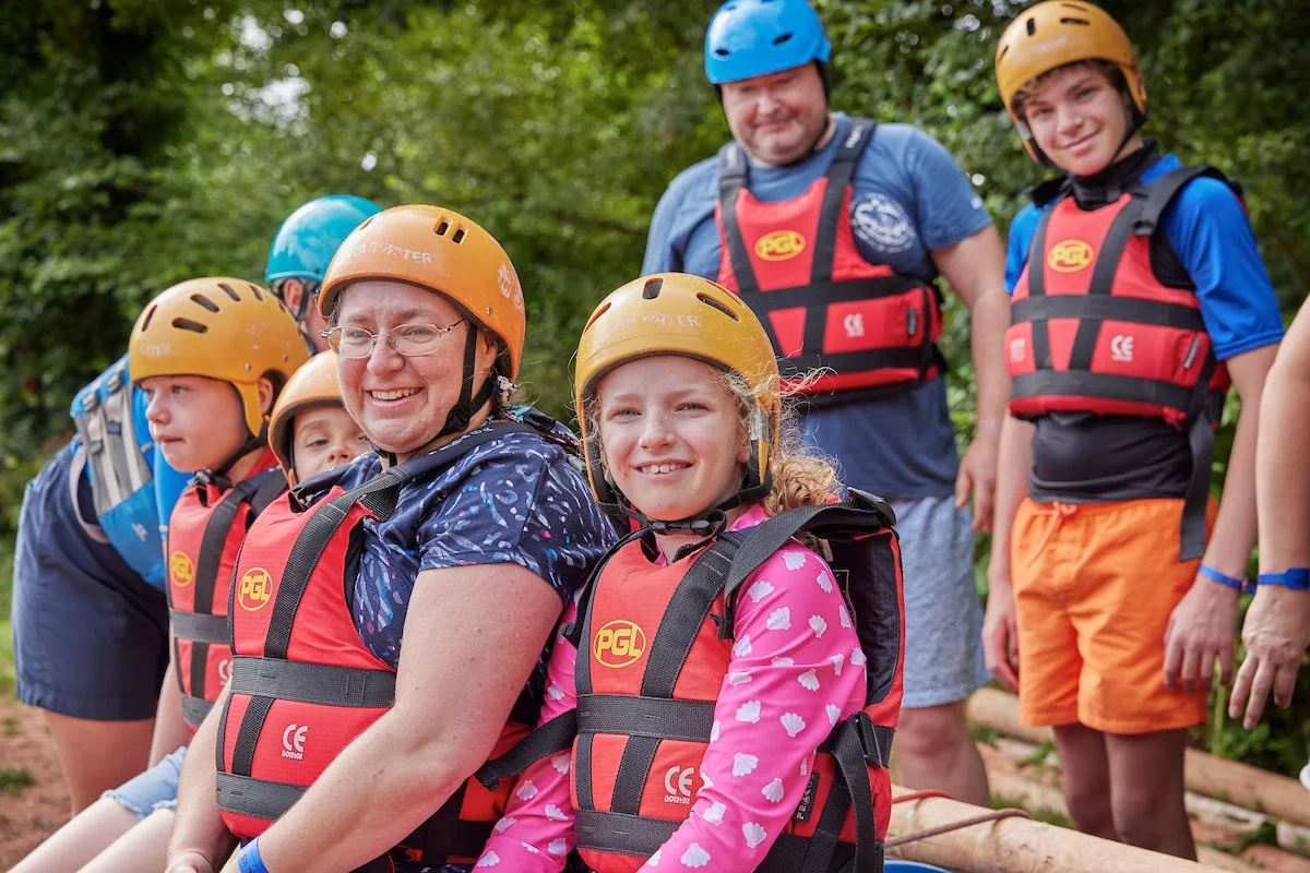 A group of mixed age pupils wearing  red buoyancy aids and orange helmets sit with two adults outside on rafts made from wood, rope and barrels..