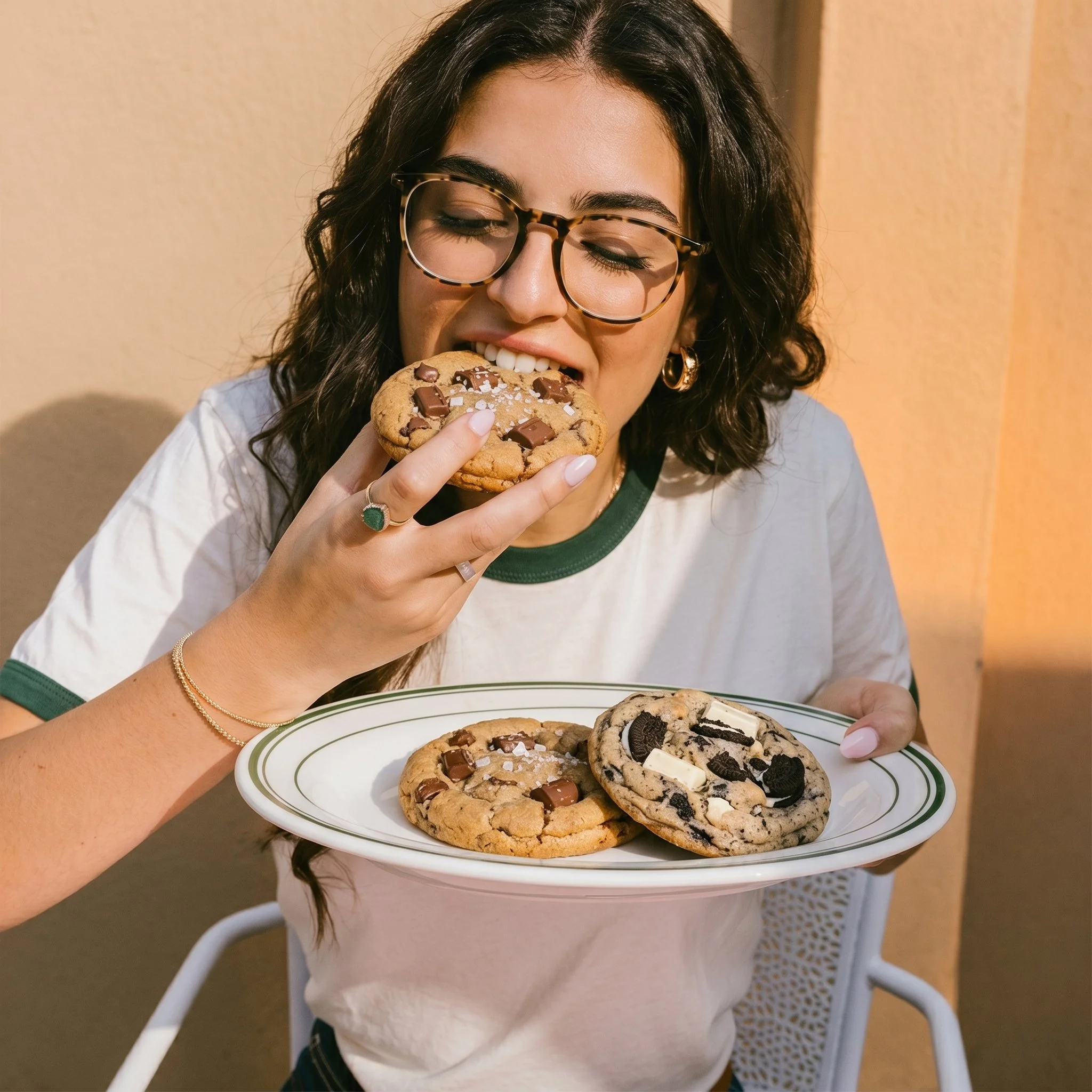 A woman with dark wavy hair, glasses, and gold jewelry is holding a plate with cookies, including chocolate chip cookies and an Oreo, as she prepares to take a bite.