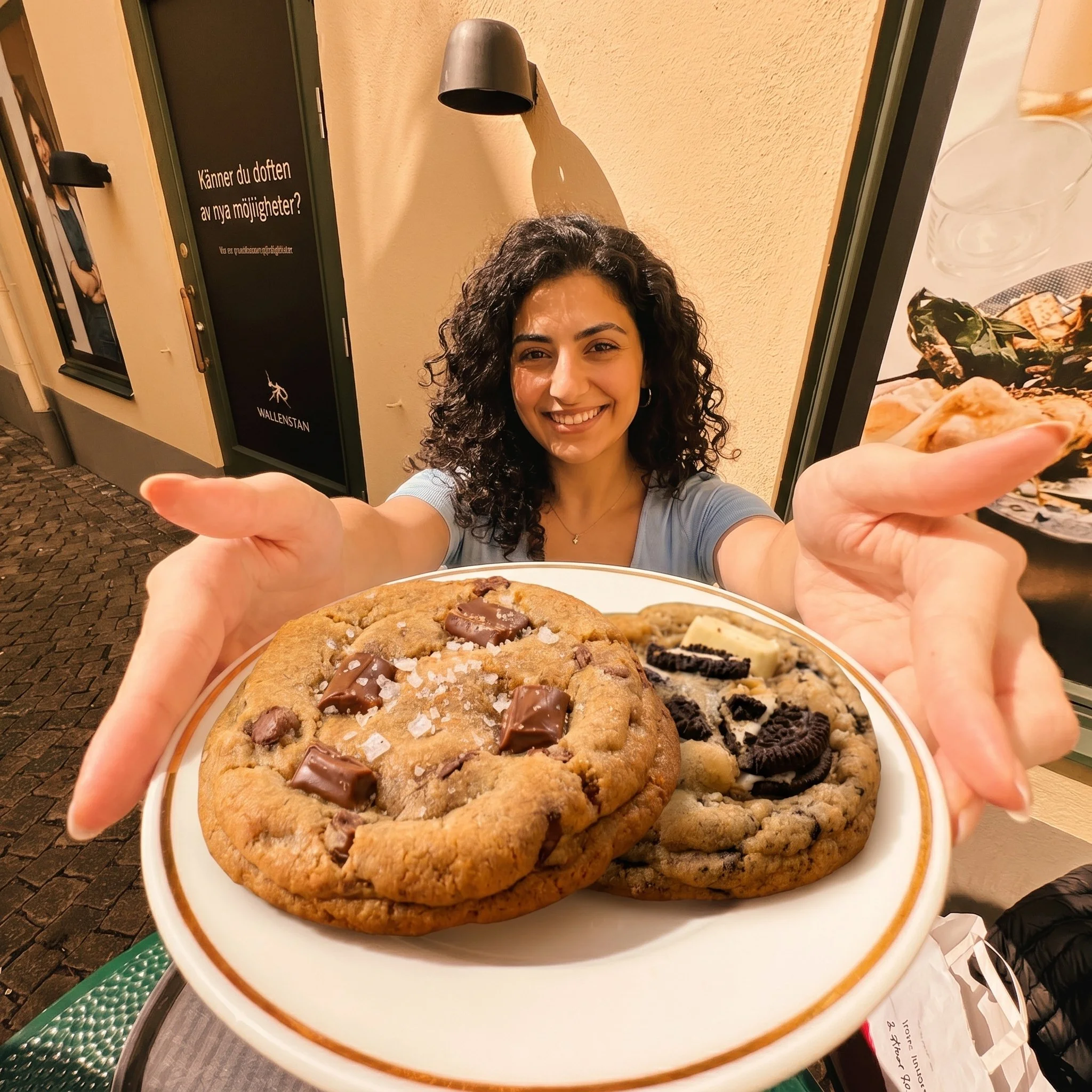 A woman smiling and taking a selfie while holding a large plate of assorted cookies, including chocolate chip and Oreo cookies, in front of a restaurant window.