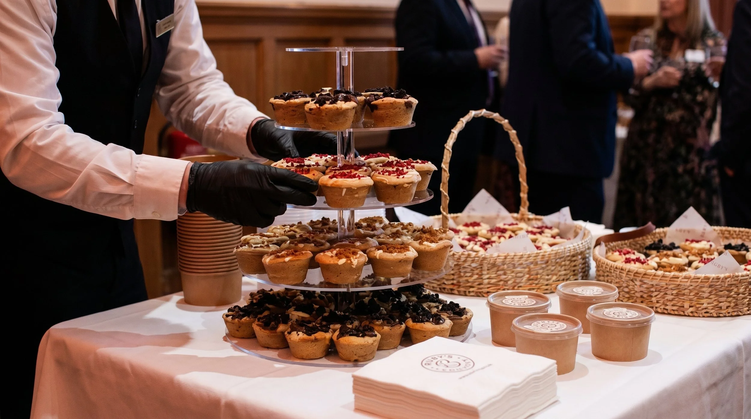 Display of assorted cupcakes arranged on a multi-tiered stand at a food event, with people in formal attire in the background.