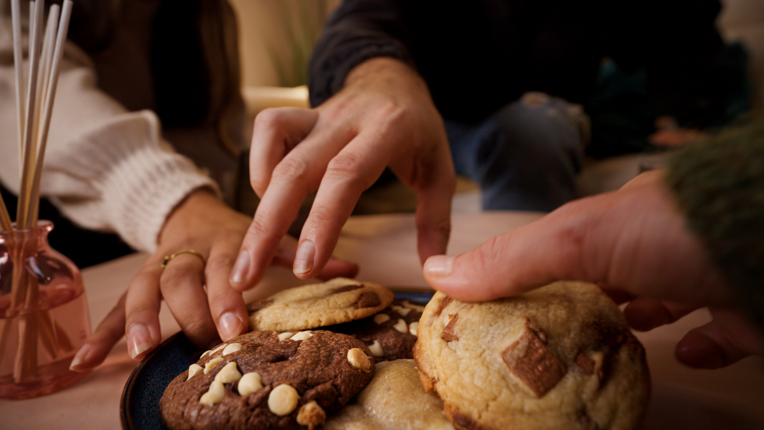 People reaching for freshly baked cookies on a platter, with a small pink vase with sticks on the side.