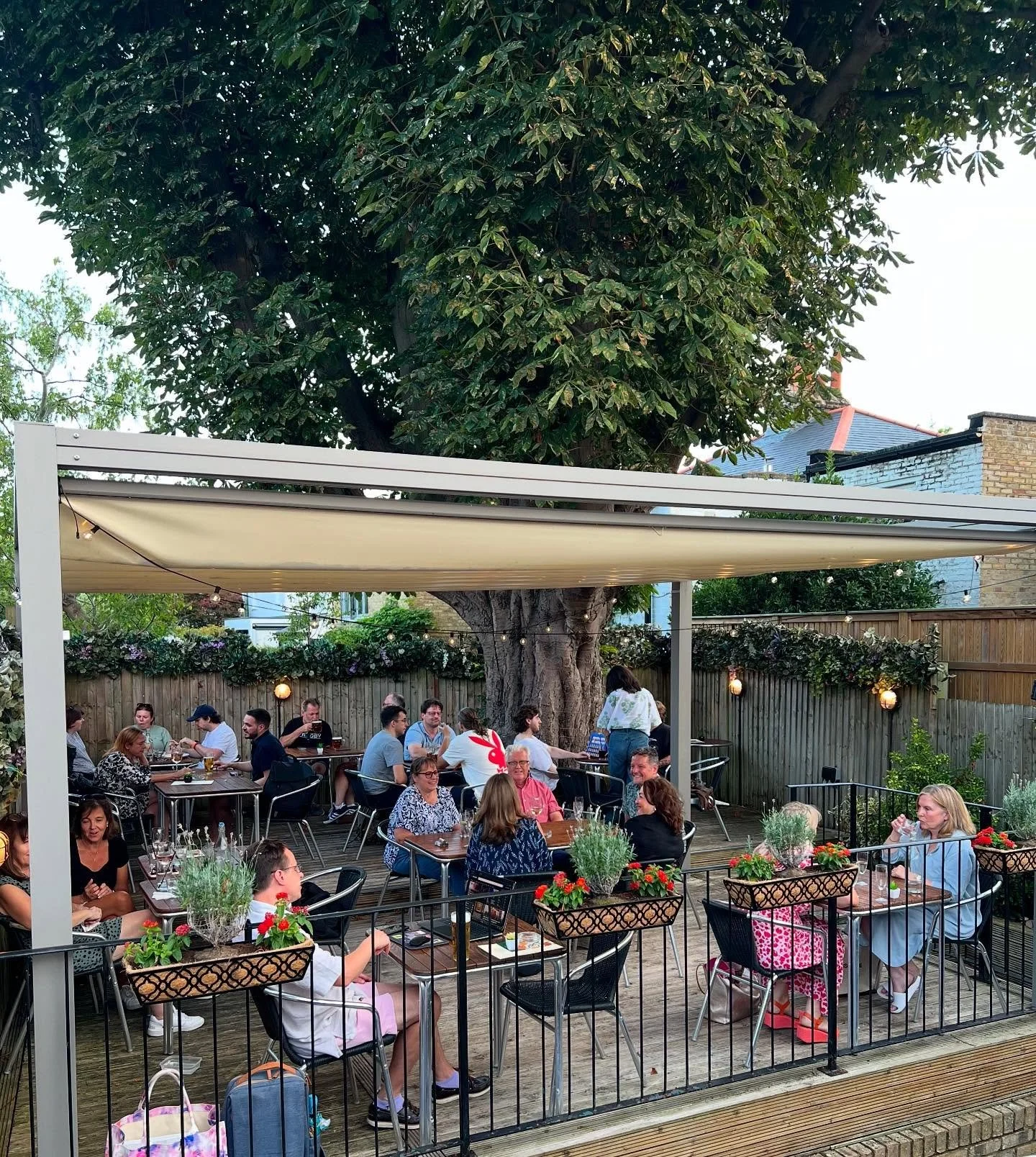 The secret garden at The Arab Boyt with people dining outdoors on a wooden deck with potted plants, surrounded by a wooden fence with hanging lights and a large leafy tree in the background.