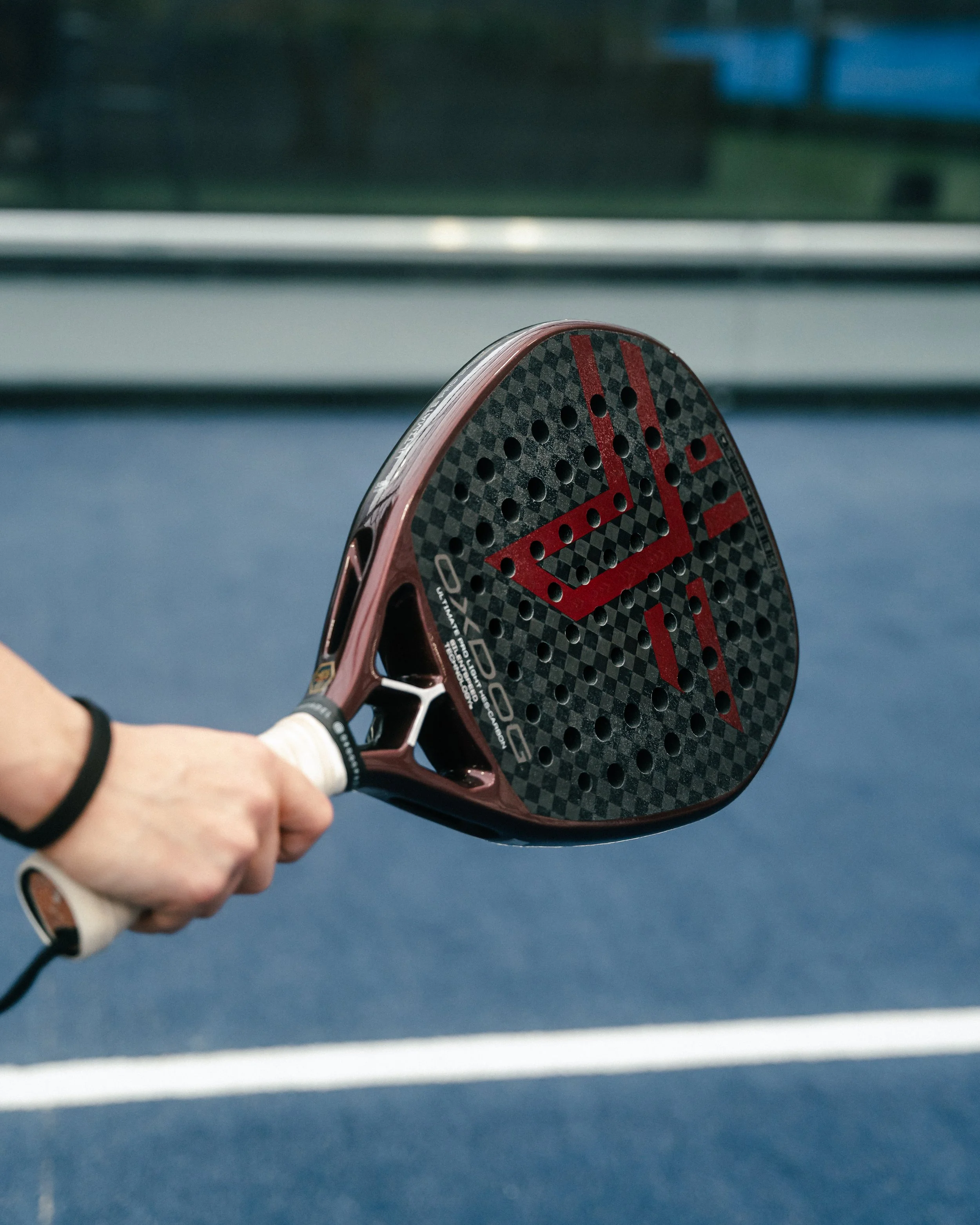 A person holding a padel racket on a court, with a blue surface and a fence in the background.