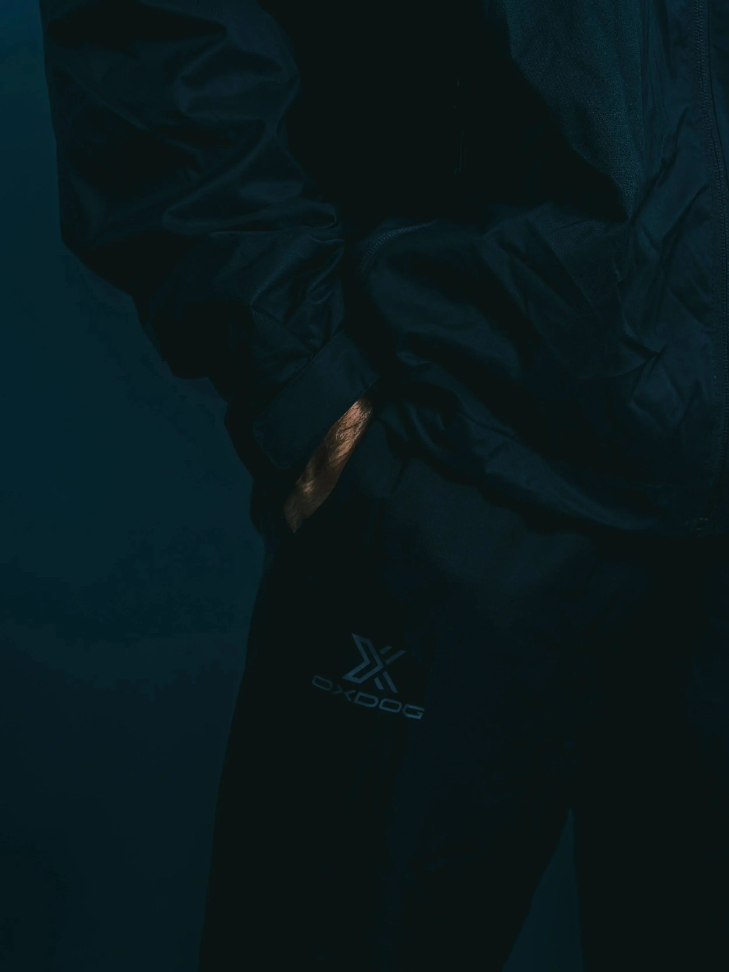 Person wearing a black jacket and black pants with a 'DOGO' logo on the pants, standing against a dark background.