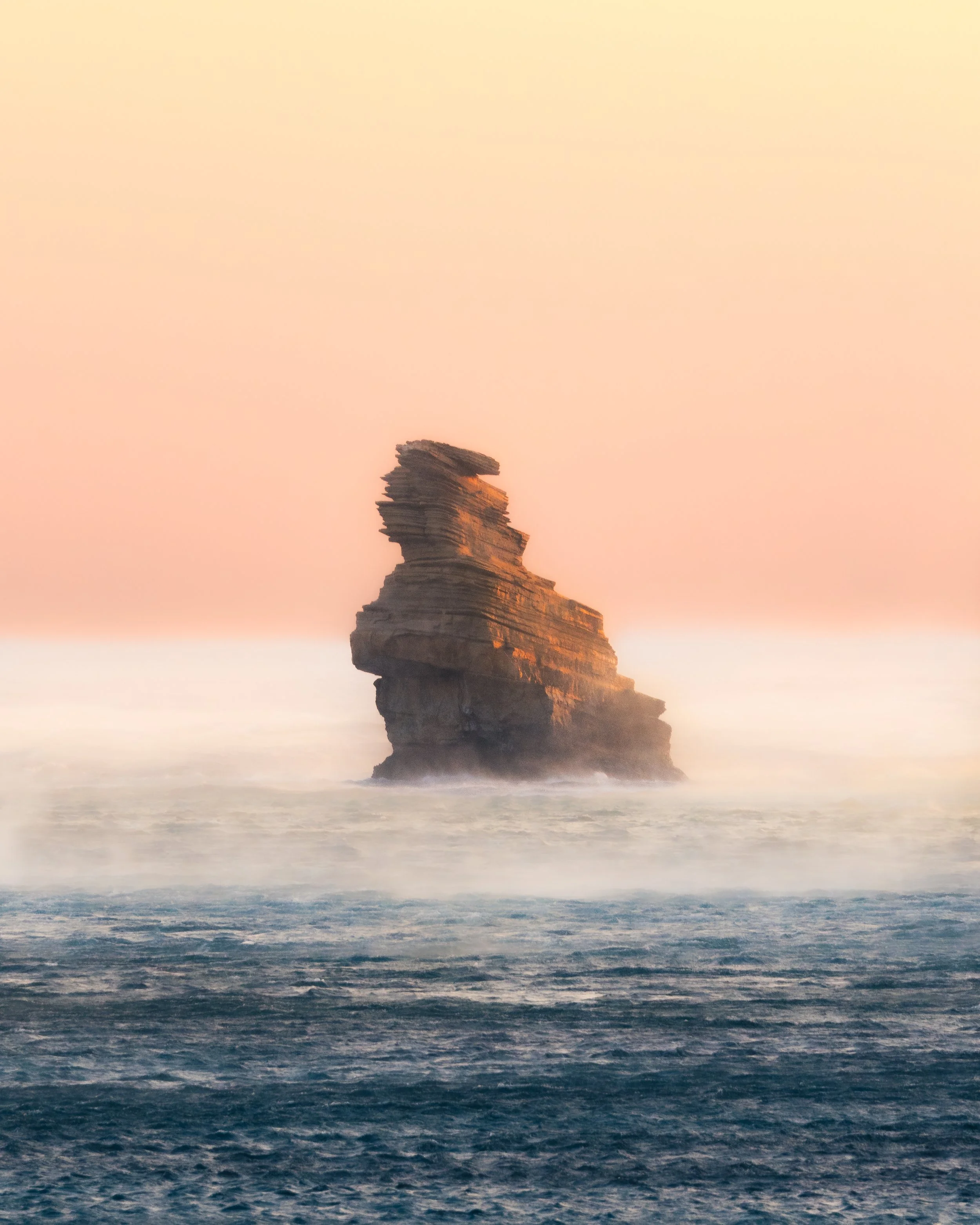 A sea stack in warm sunset light above calm waves.