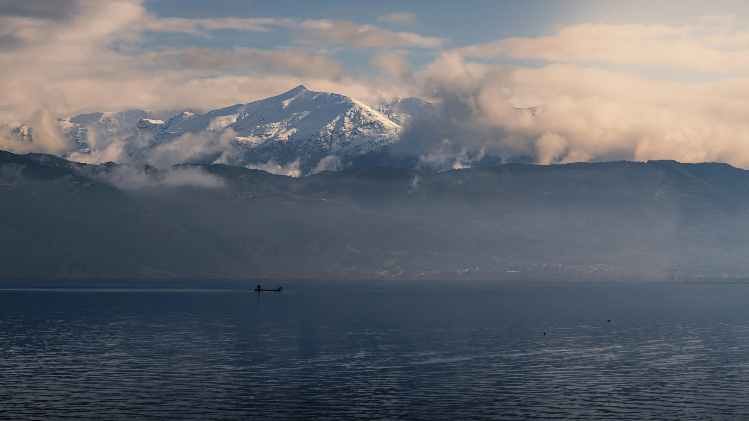 Snow-capped mountains behind a large body of water with a small boat and a person in the boat.