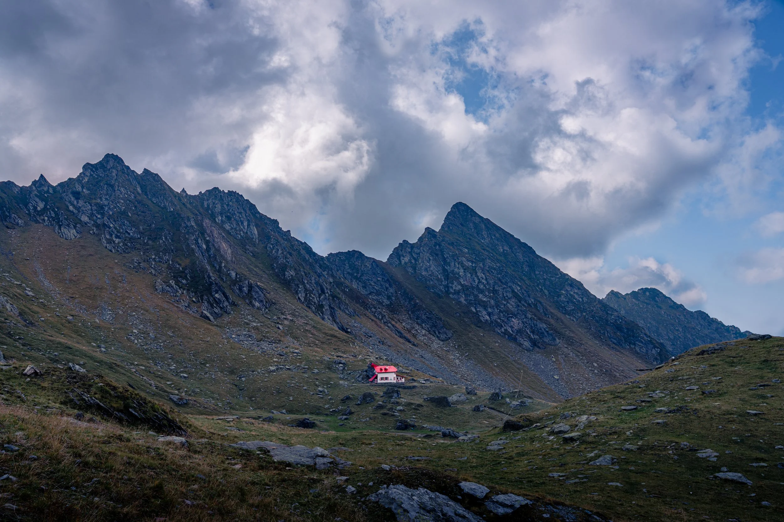 A small house with a red roof and white walls situated in a green valley at the base of rocky, jagged mountain peaks under a sky filled with clouds.