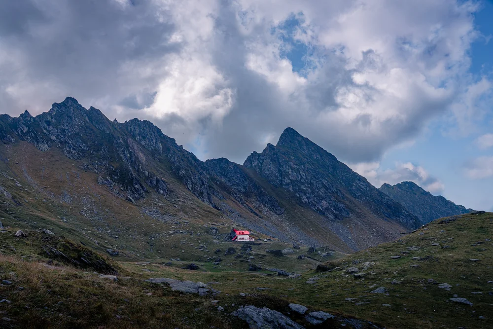 A small red-roofed house in a green valley beneath jagged mountain peaks.