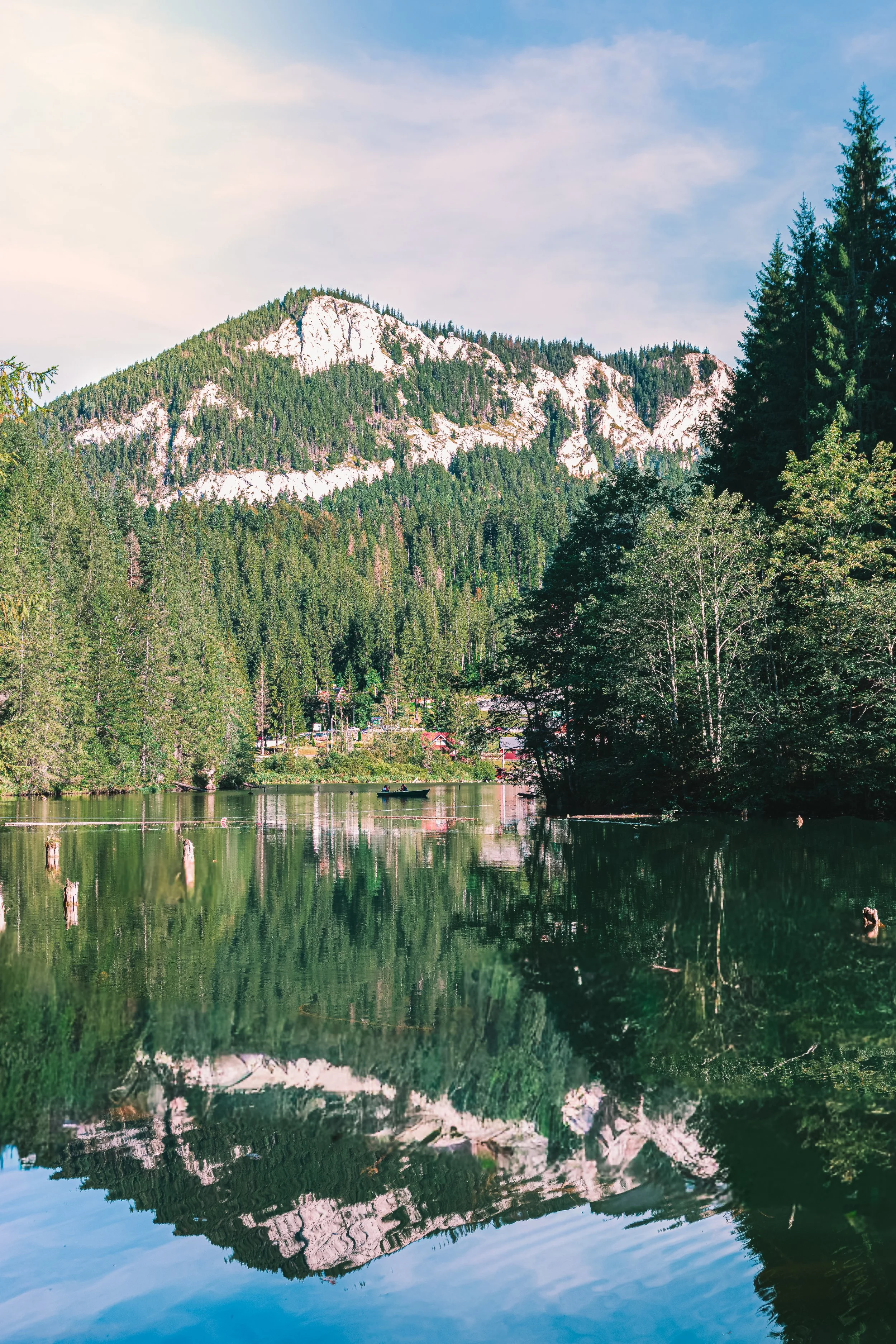 Scenic view of a mountain with rocky cliffs and dense green forest, reflected in a calm river with boats and trees.