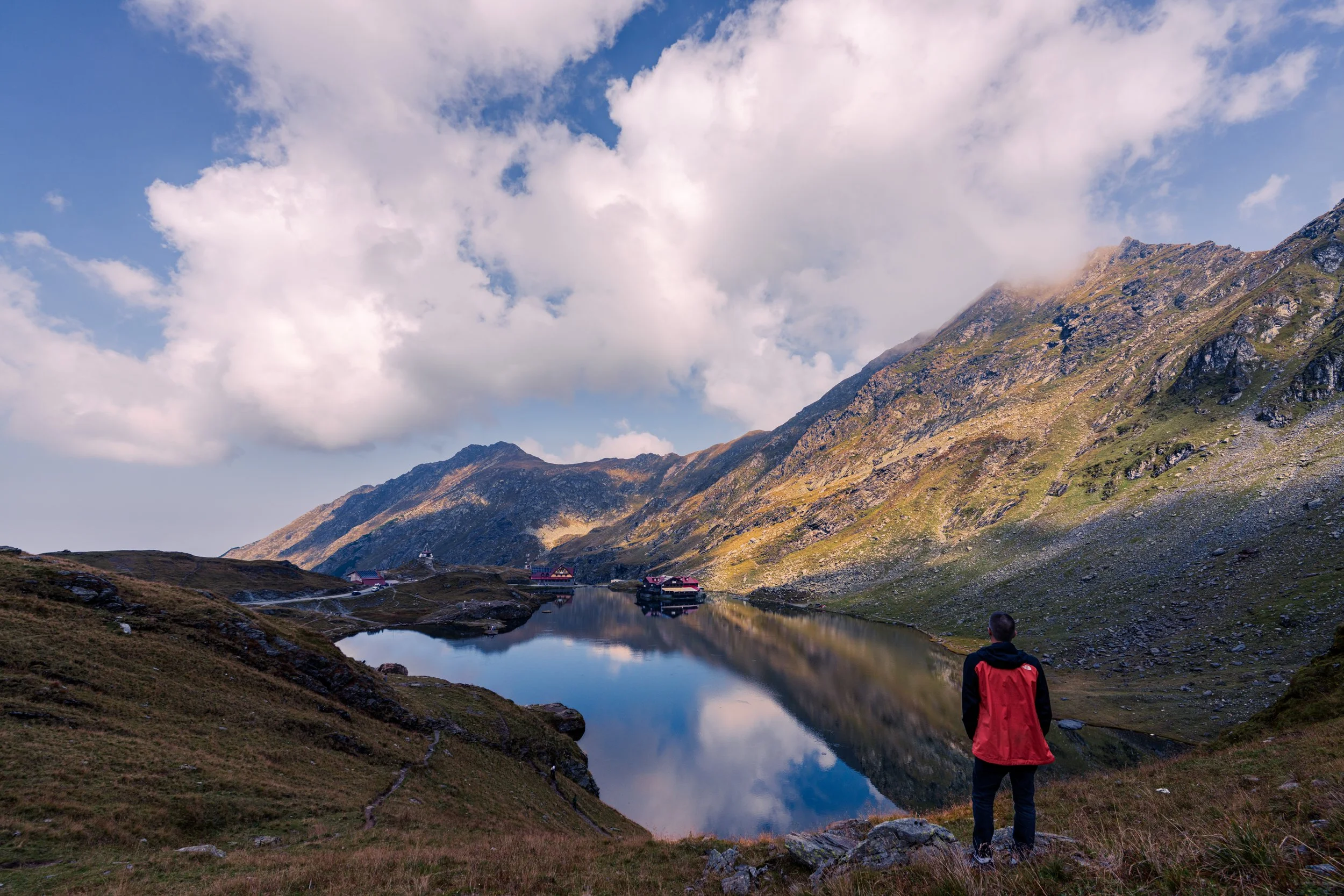 A person wearing a red jacket and black pants standing on a grassy hillside overlooking a mountain lake with reflections of the sky and clouds, surrounded by rugged mountains with a small cluster of buildings near the water.