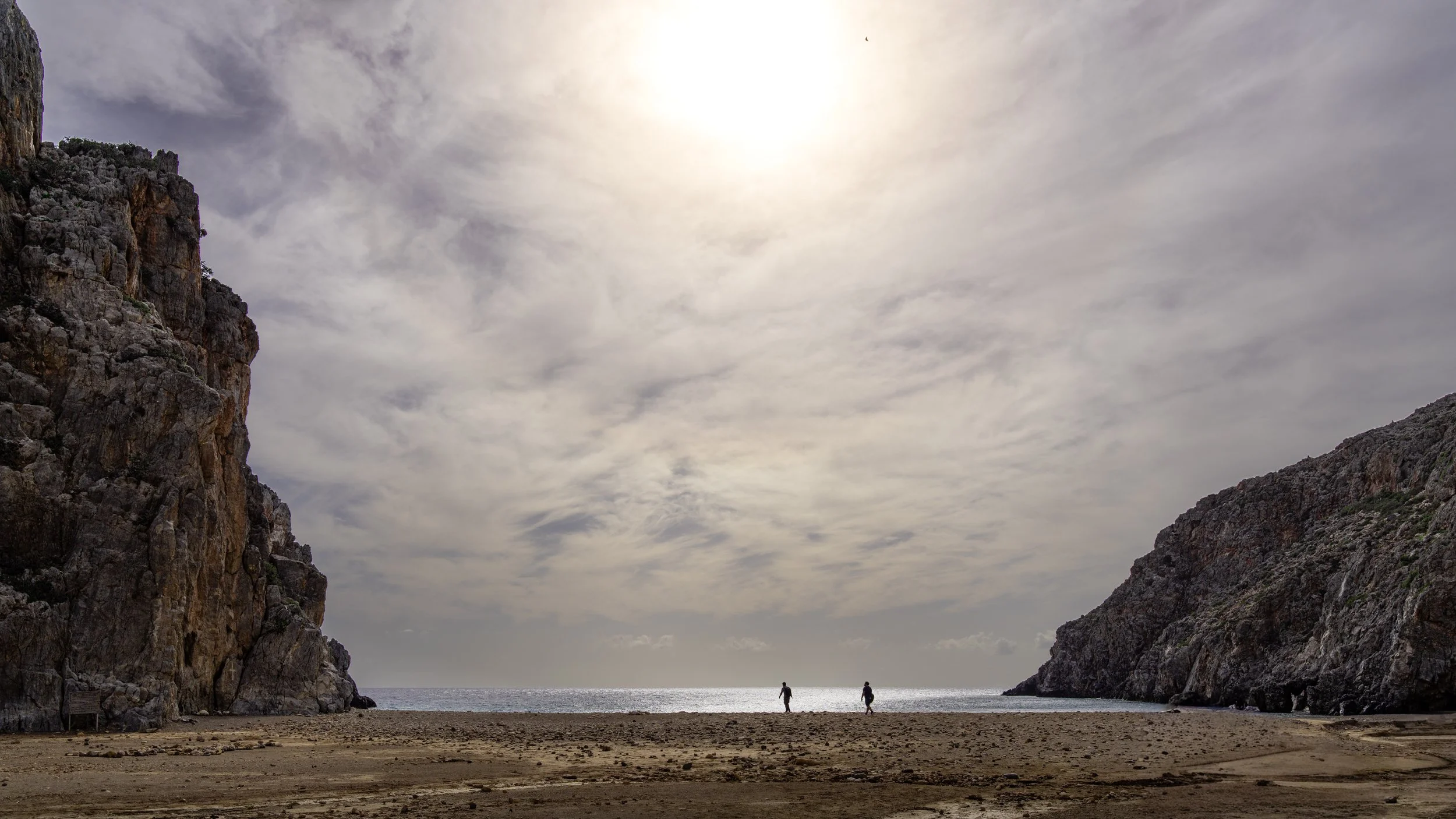 A beach scene with two people walking along the shoreline between tall rocky cliffs under a cloudy sky with the sun partially visible.