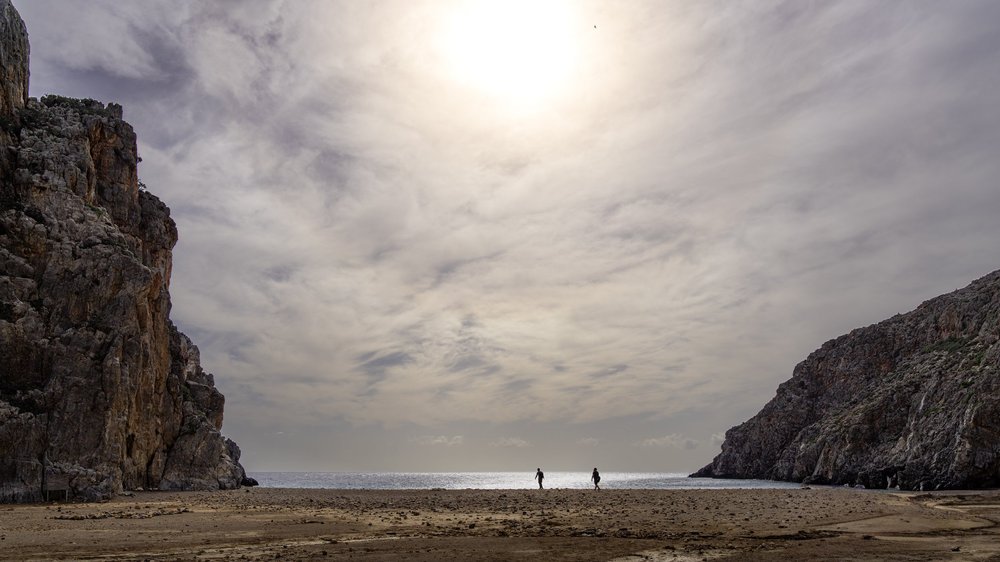 Two people walking along a beach between tall cliffs under a cloudy sky.