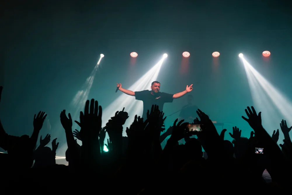 Anser with arms outstretched on stage above raised hands in the audience.