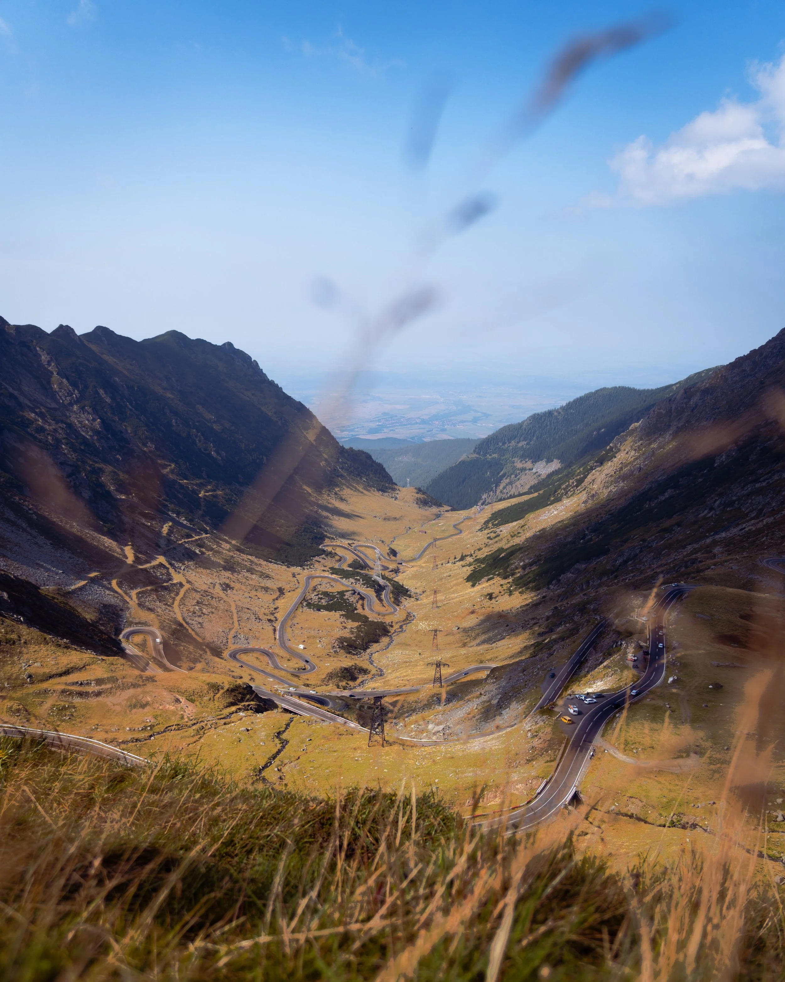 Mountain valley with winding roads through grassy slopes, distant mountains under a clear blue sky with some clouds, viewed from a high vantage point.