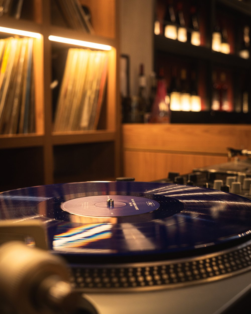 A record on a turntable beside shelves of vinyl and wine.