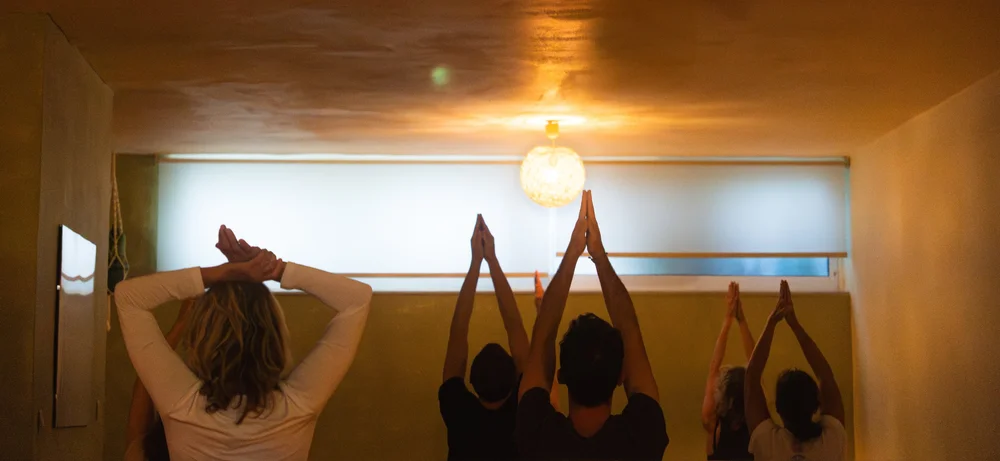 Yoga participants stretching their arms overhead in a softly lit studio.