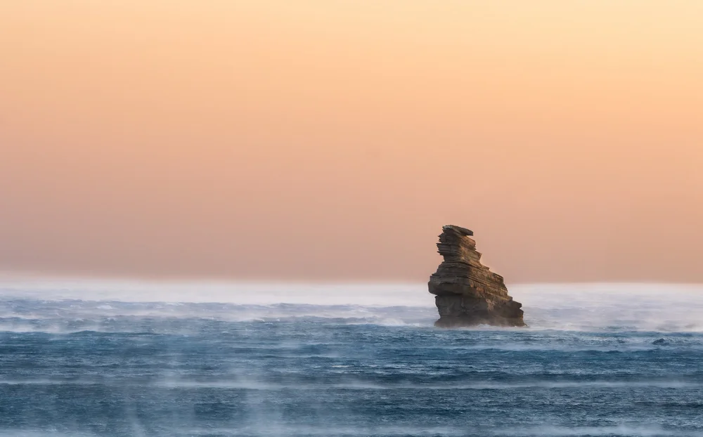 A sea stack above gentle waves beneath an orange-pink sunset sky.