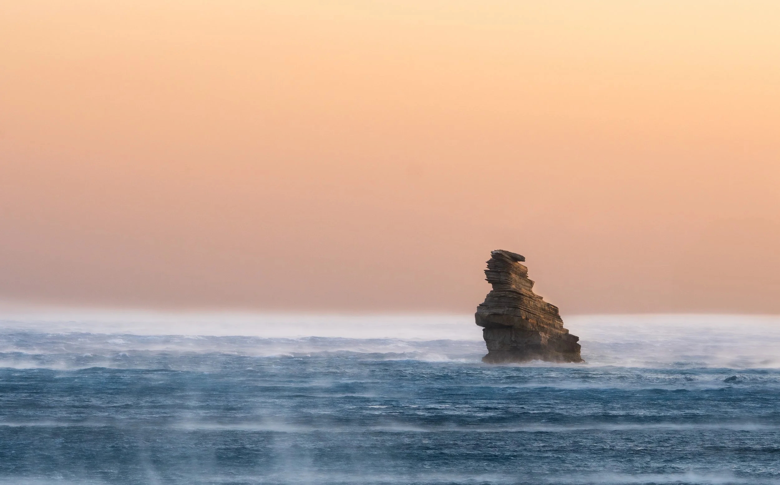 A sea stack above gentle waves beneath an orange-pink sunset sky.