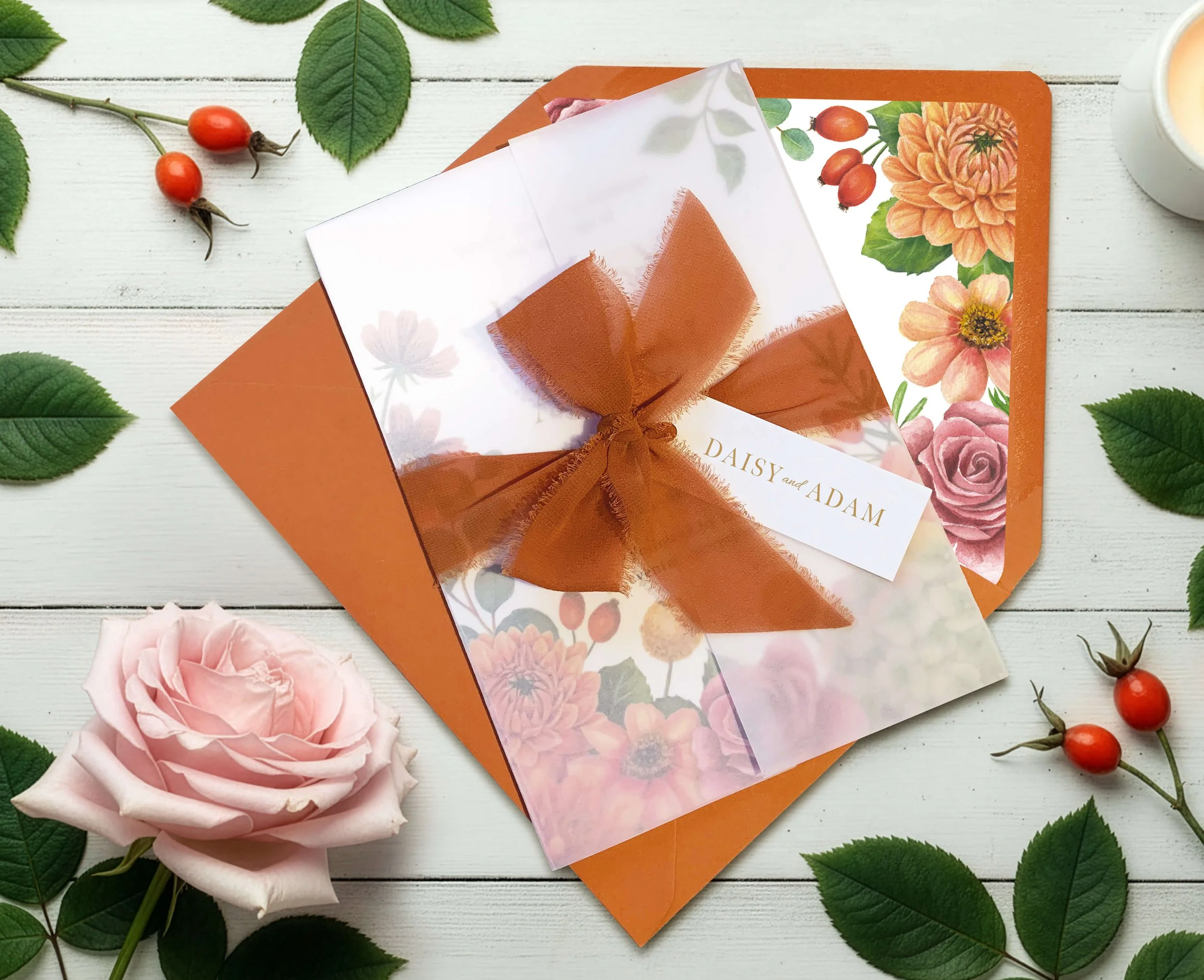 A floral wedding invitation with a burnt orange ribbon bow, placed on a white wooden surface with roses, green leaves, rose hips, and a partially visible cup of tea or coffee.