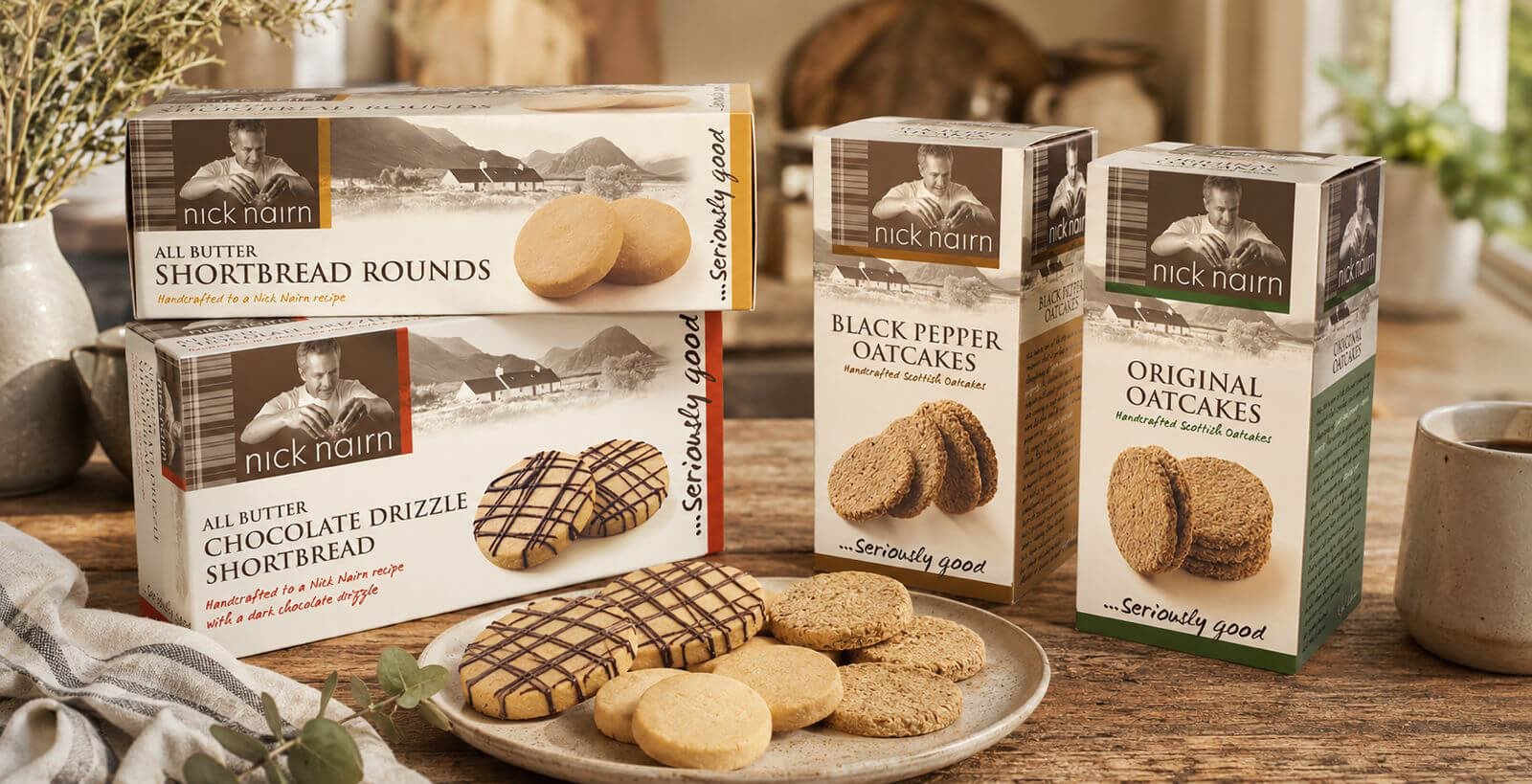 A selection of Nick Nairn oatcakes and shortbread cookies on a wooden table, with boxes of original oatcakes, black pepper oatcakes, chocolate drizzle shortbread, and all butter shortbread rounds.