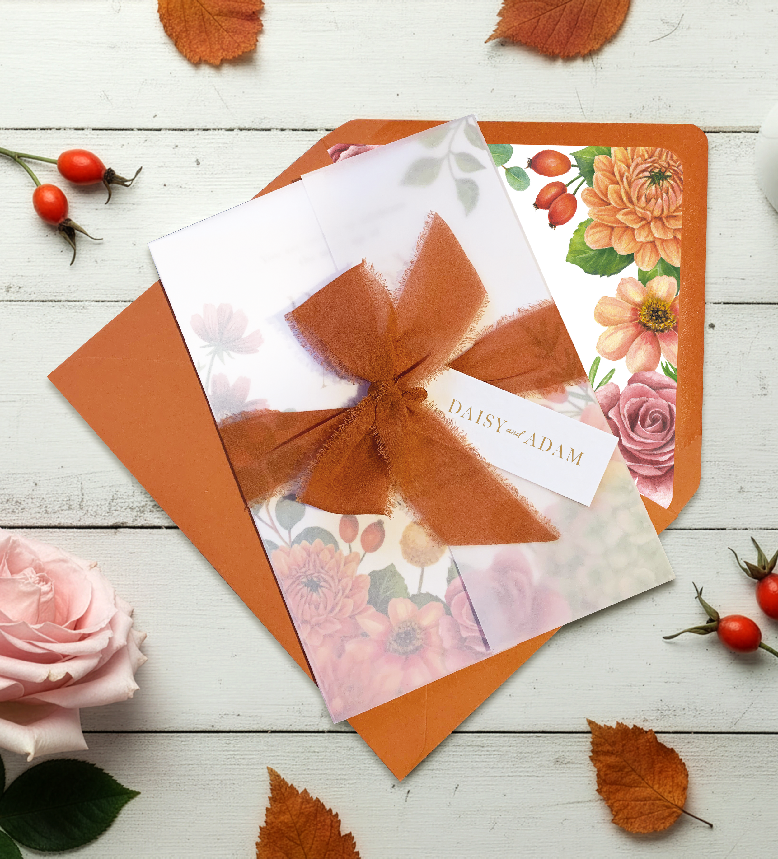 A floral invitation card with a brown ribbon bow, placed on a white wooden surface surrounded by scattered orange autumn leaves, pink roses, and small red berries.