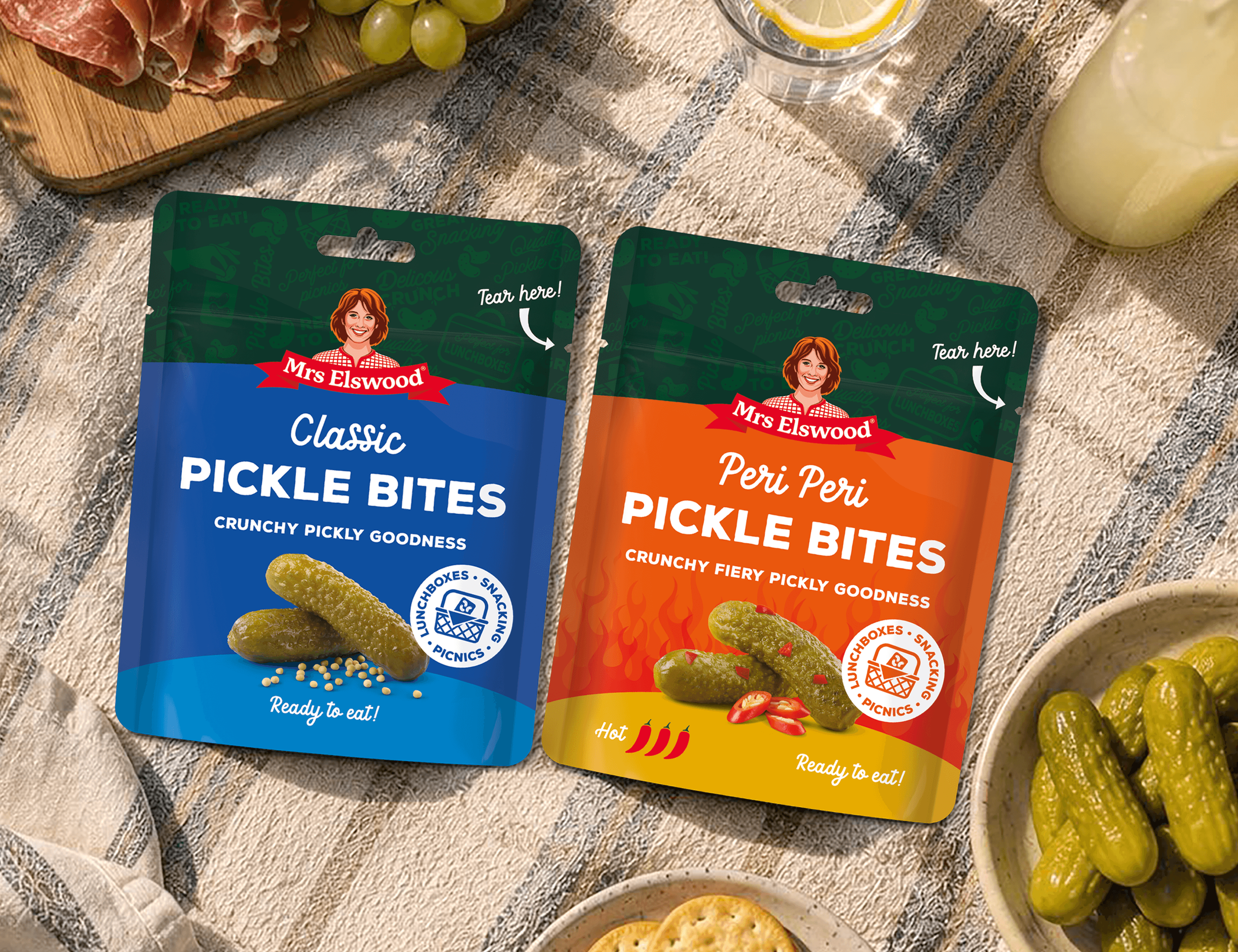 Two packages of Mrs Elswood pickle bites, one blue labeled 'Classic' and one orange labeled 'Peri Peri,' placed on a textured beige surface with grapes, a glass of lemon water, and a bowl of pickles nearby.