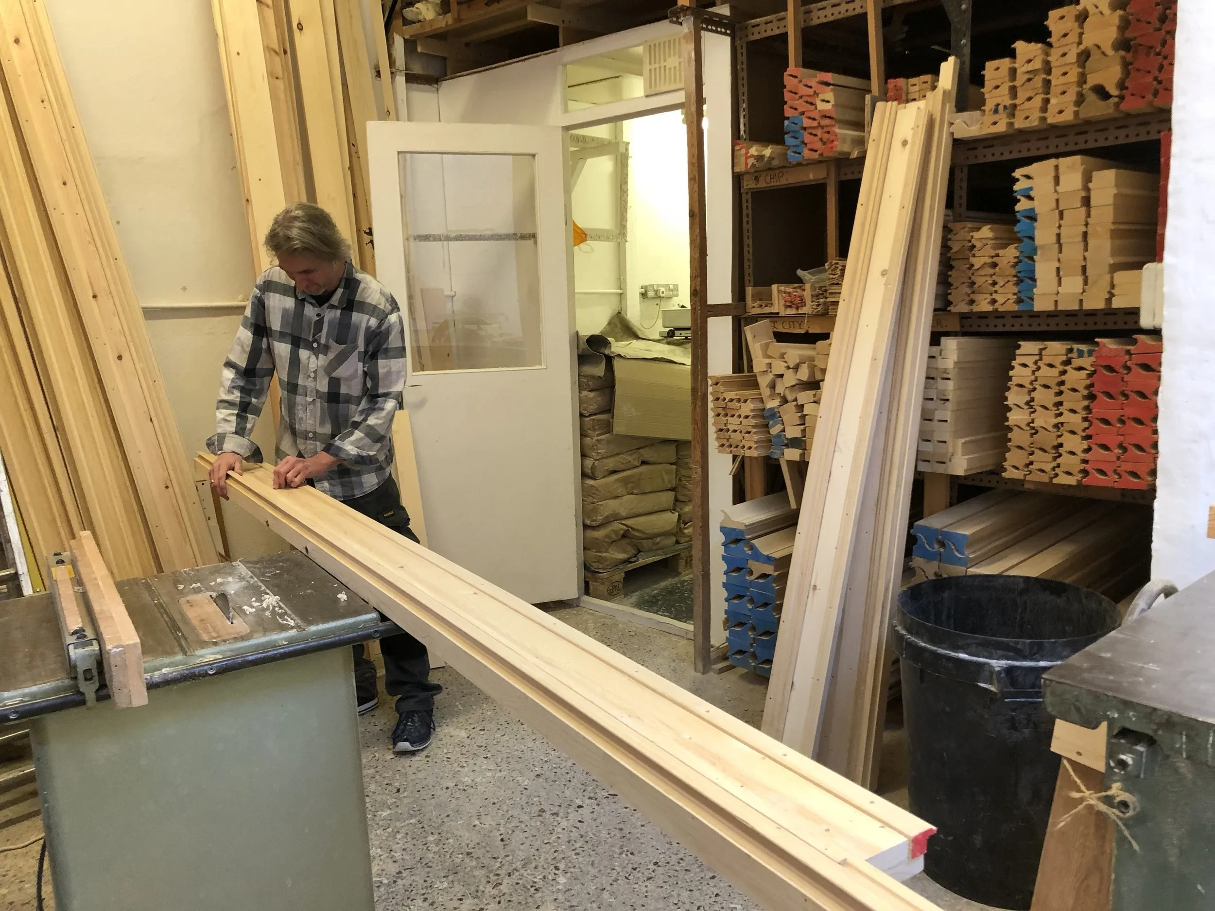 A person wearing a plaid shirt looks down while working with long wooden planks in a woodworking shop. Shelves behind are filled with stacked wood pieces of various shapes and colors. The workspace is organized with tools and materials.