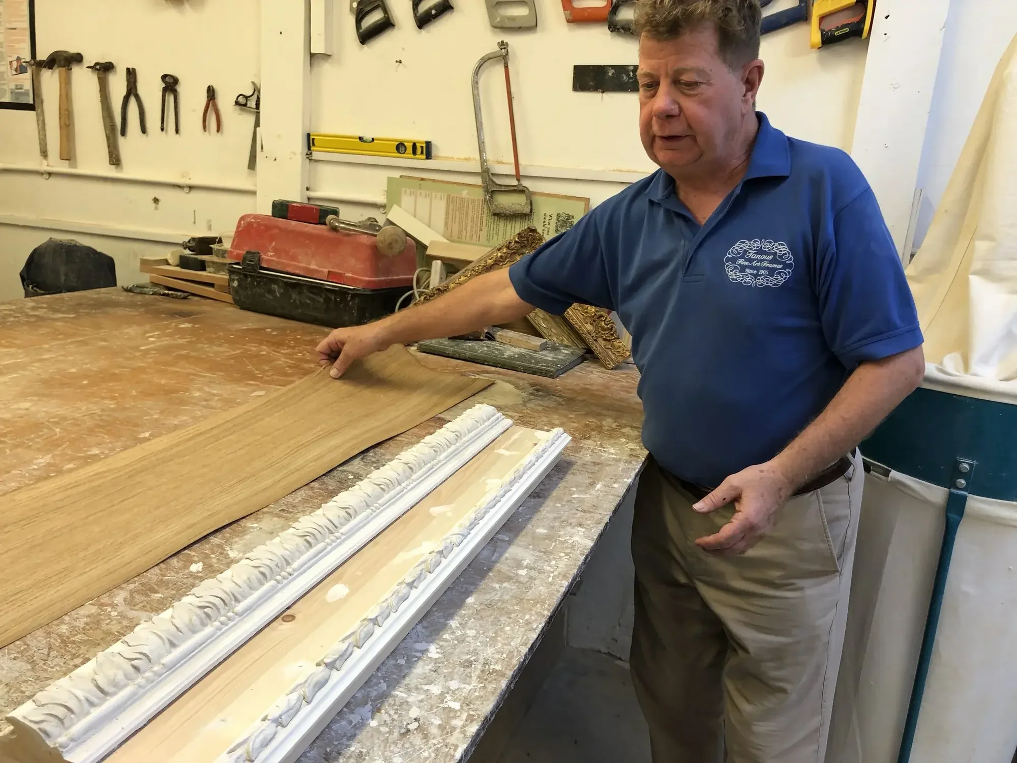 A man working in a workshop or studio, sitting at a workbench, pointing to a decorative molding or trim piece. The background features tools such as hammers, saws, and level hanging on a white wall.
