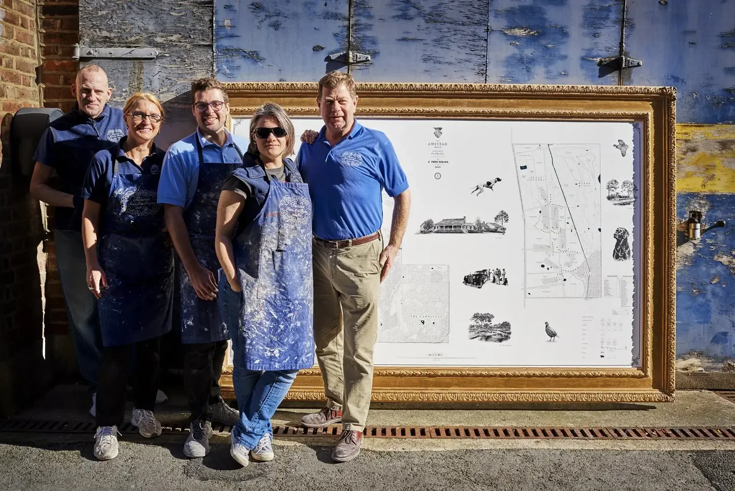 Group of five people standing outdoors in front of a large framed blueprint or map, some wearing aprons with paint stains, sunny weather, brick wall to the left, and blue painted wall with peeling paint behind.