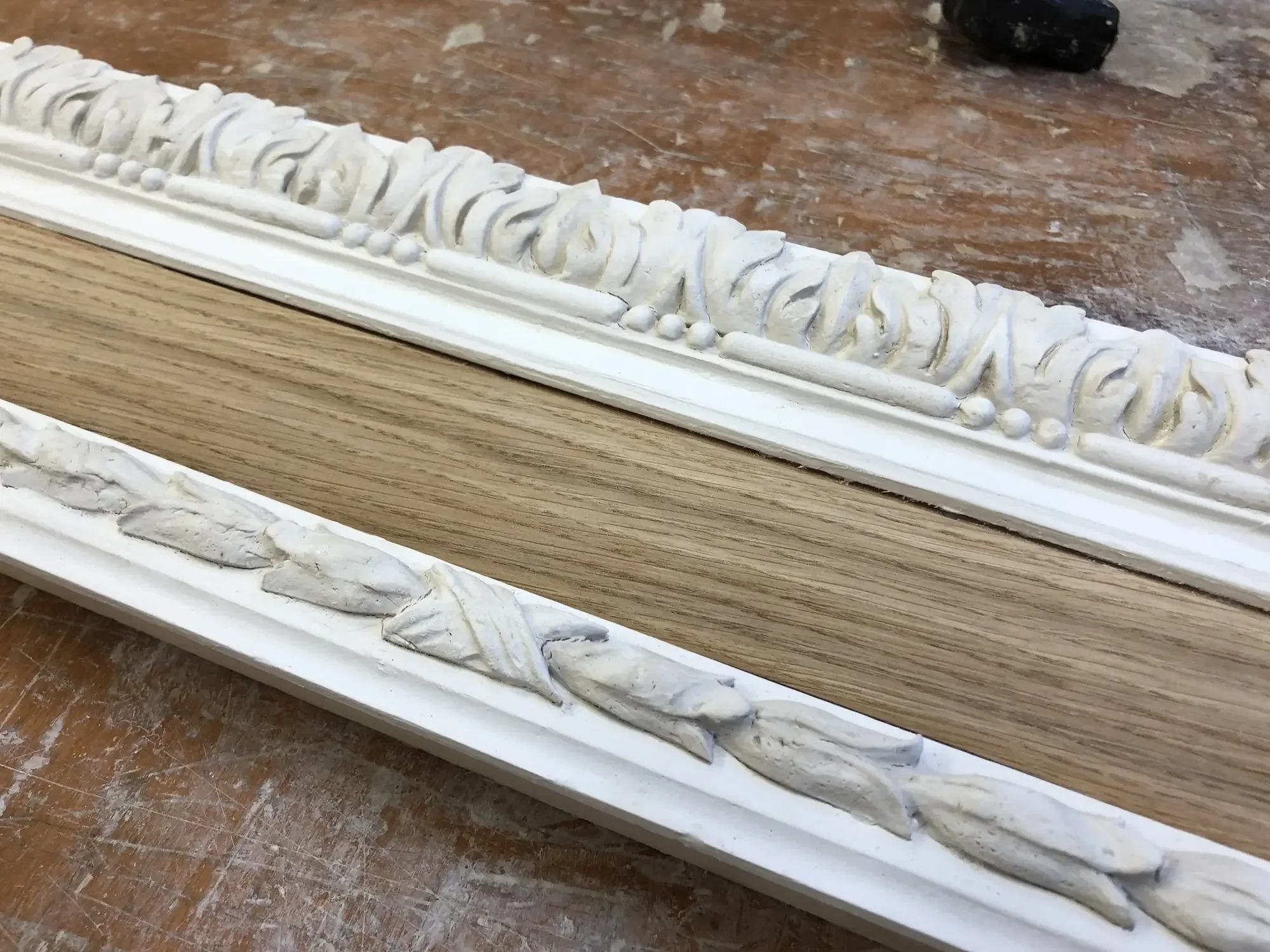 Close-up of decorative white molding with intricate scrollwork and bead details on a wooden surface, with a brownish background and a black object partially visible in the top right corner.