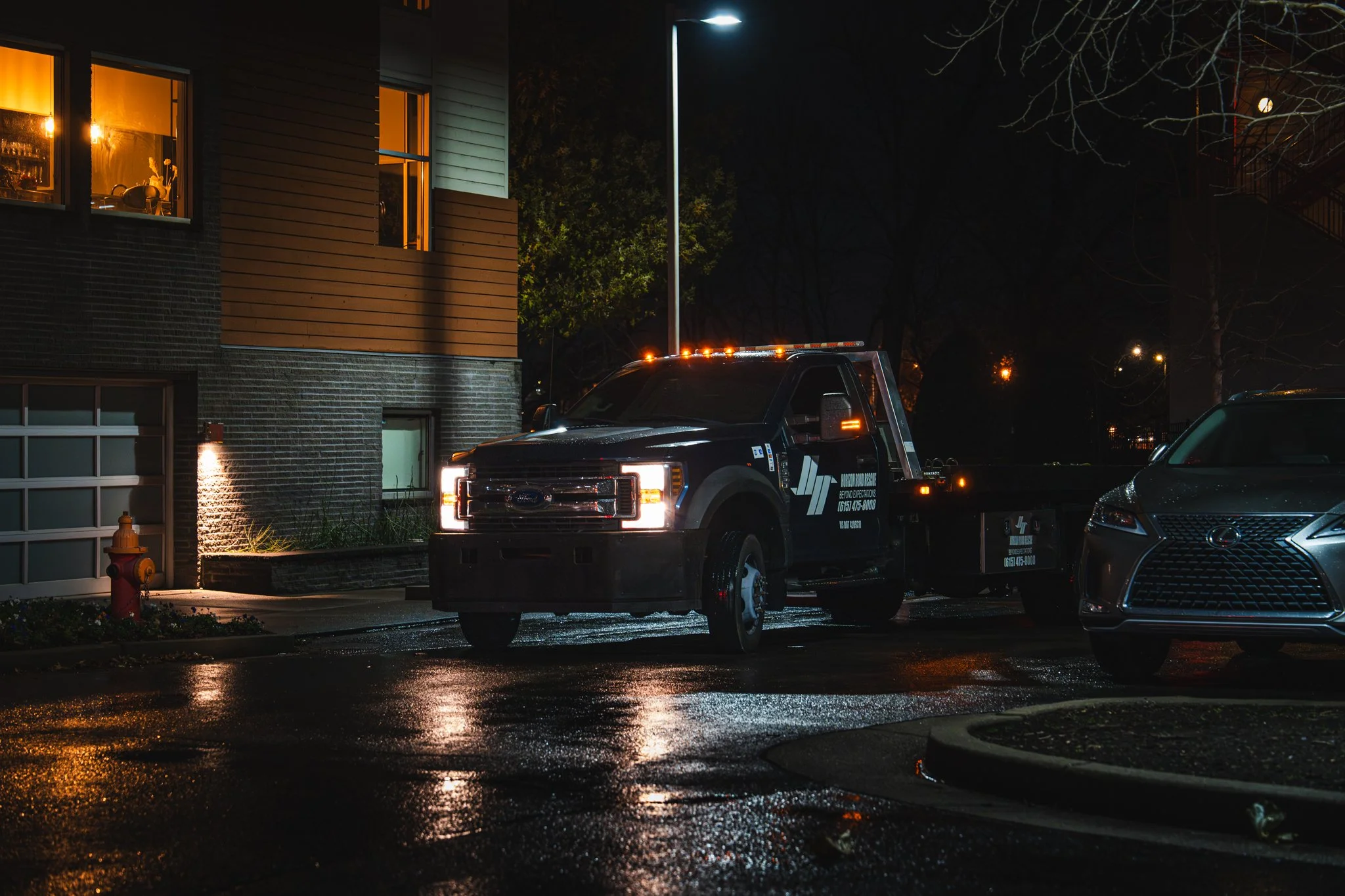 Tow truck parked at night beside a residential building, with wet pavement and streetlights illuminating the scene.