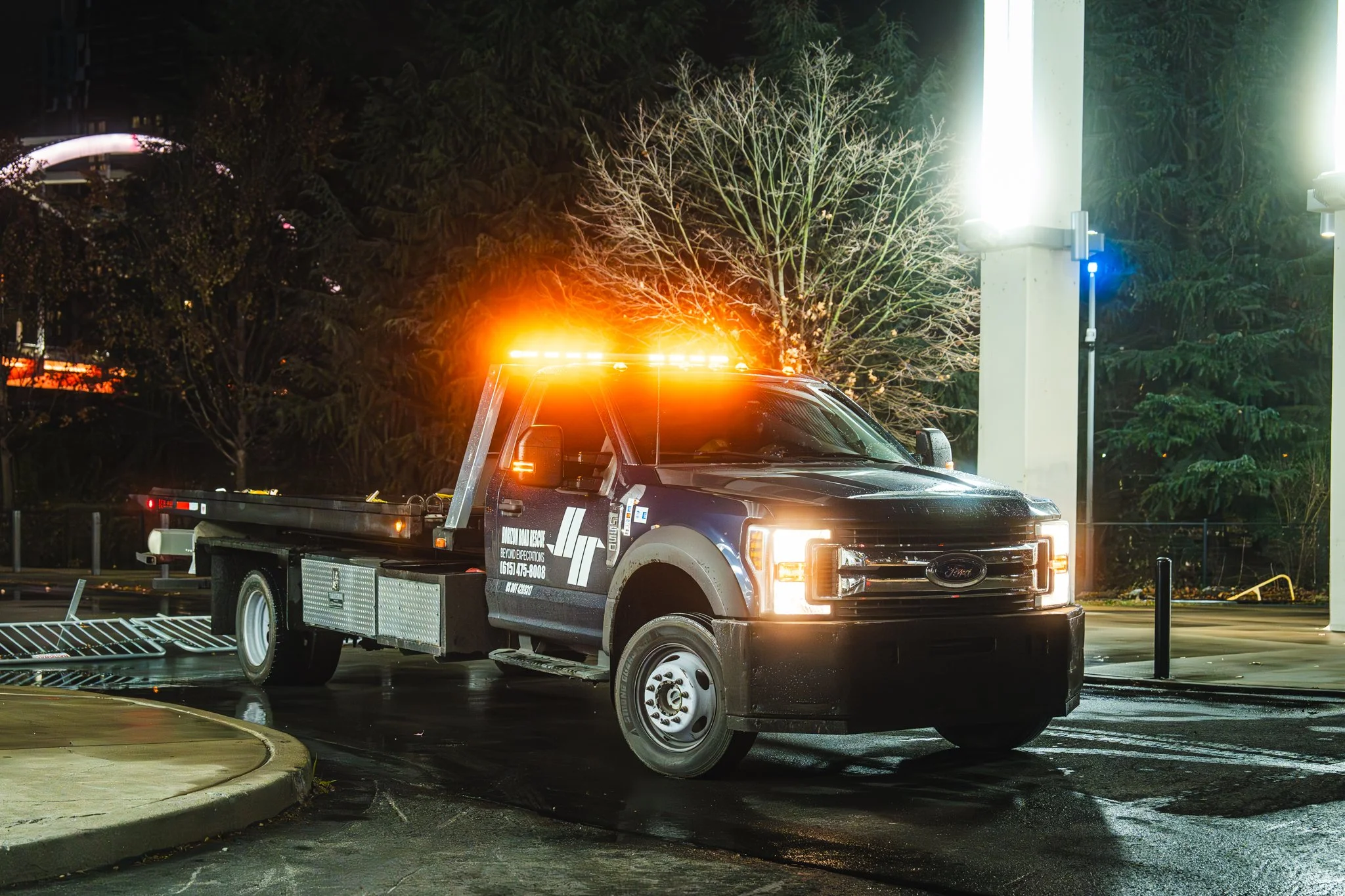 Tow truck with flashing orange lights at night in an urban setting.