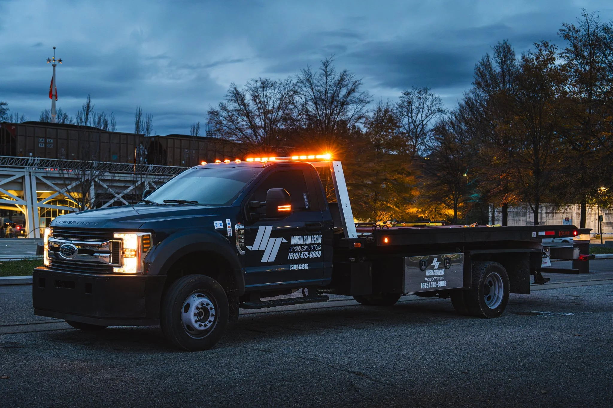 Tow truck with "Horizon Road Rescue" branding and lights on, parked near a train and trees at dusk.