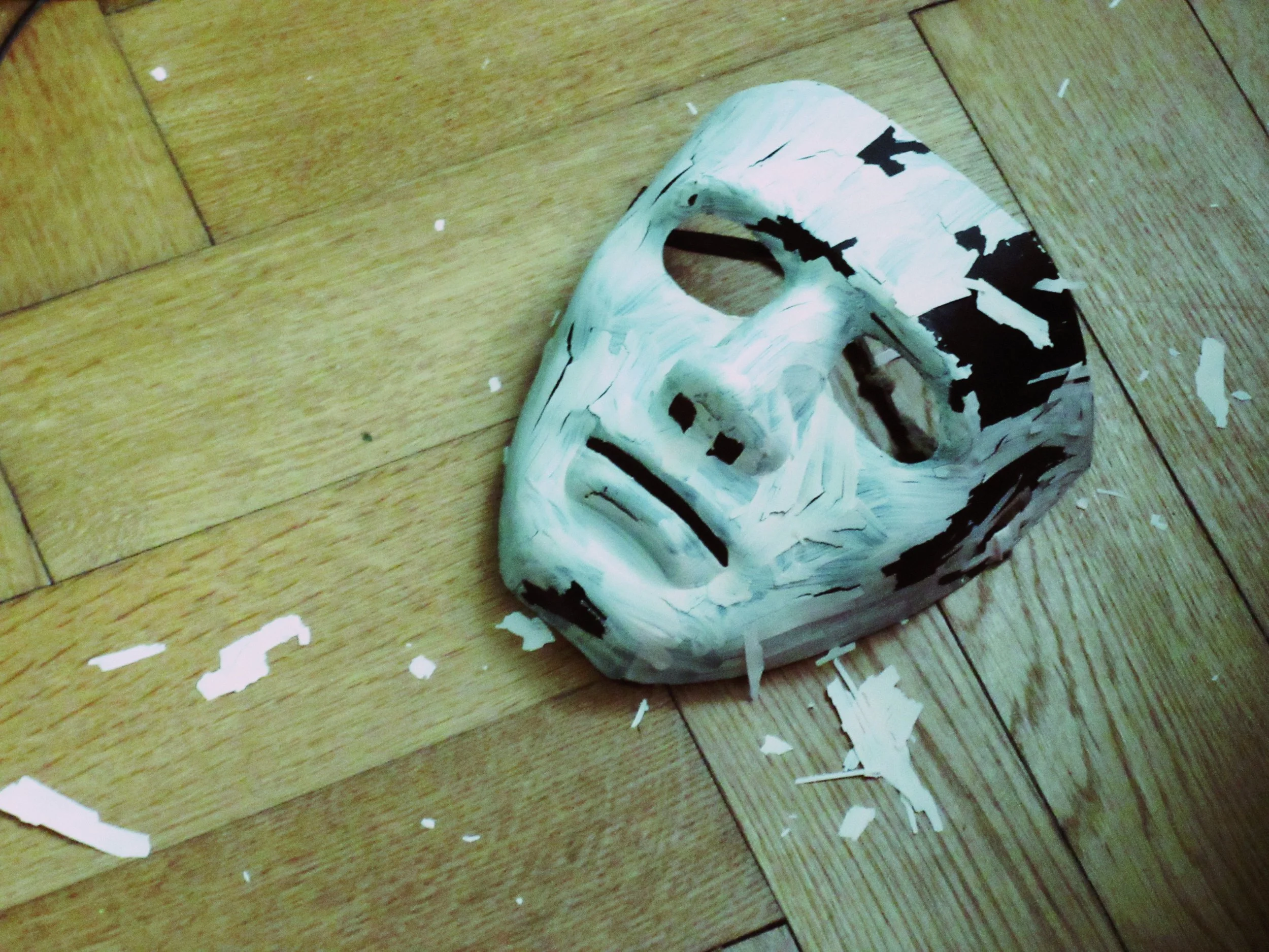 A white and black distressed mask lies on a wooden floor, creating an artistic and mysterious scene