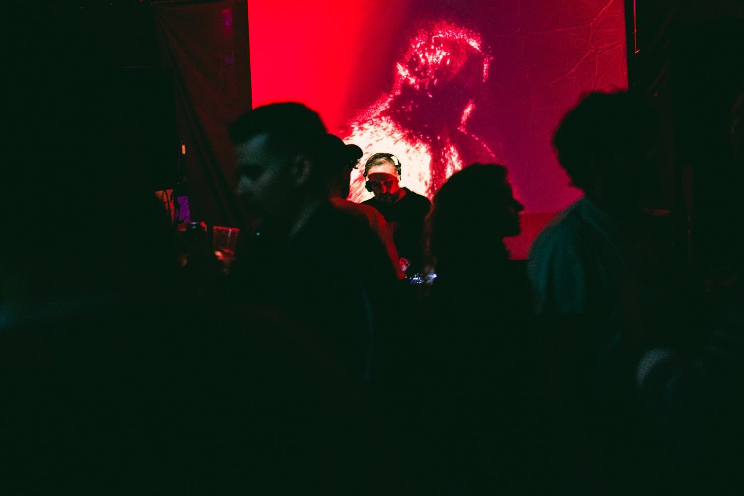 People dancing and socializing in a dimly lit nightclub with a red and pink abstract digital art backdrop.