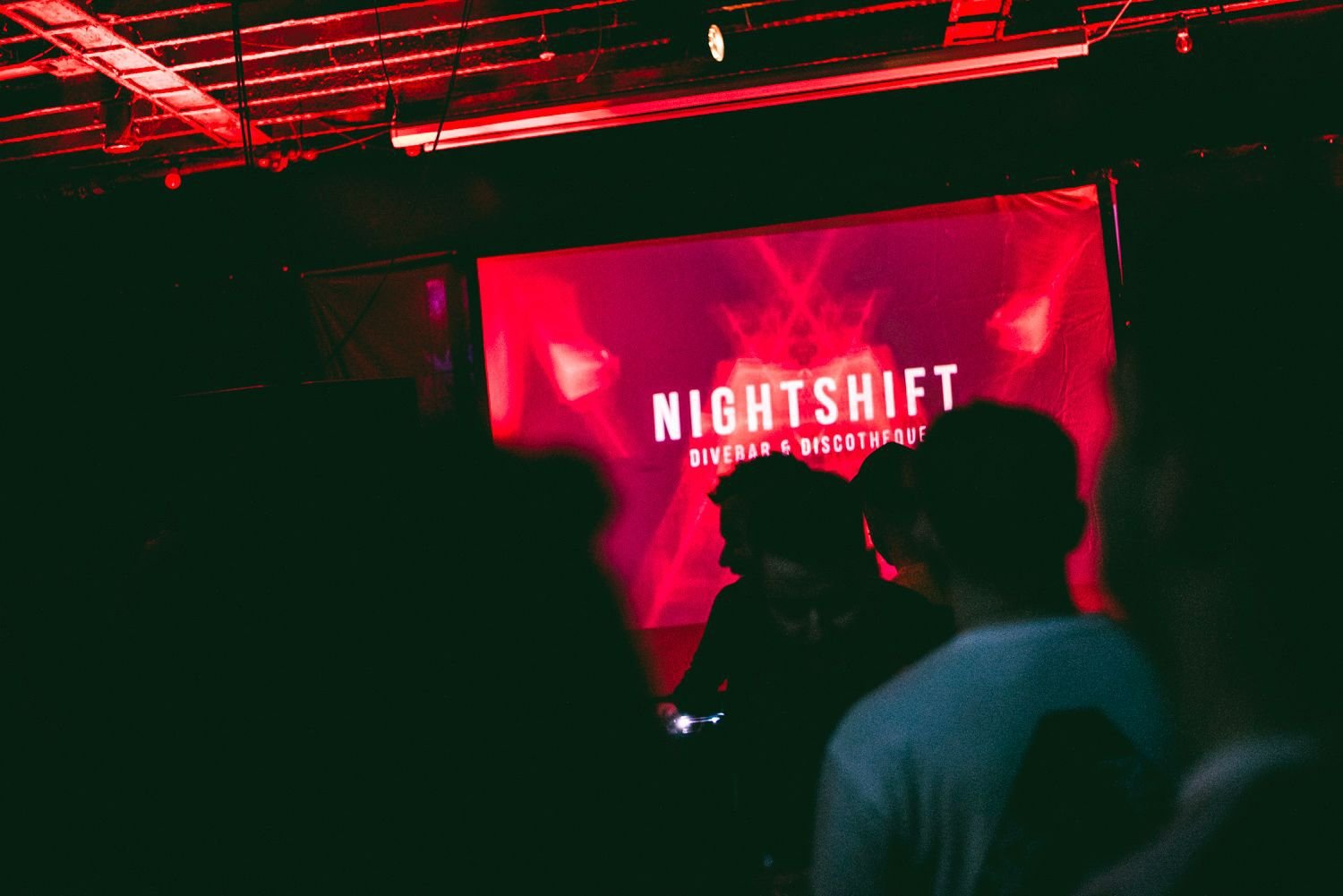 A dark indoor scene at a nightclub or bar with red lighting. A large screen displays the text 'NIGHTSHIFT' with smaller text below that reads 'DIVER BAR & DISCOTHEQUE.' Silhouettes of people are visible in the foreground, some facing towards the screen.