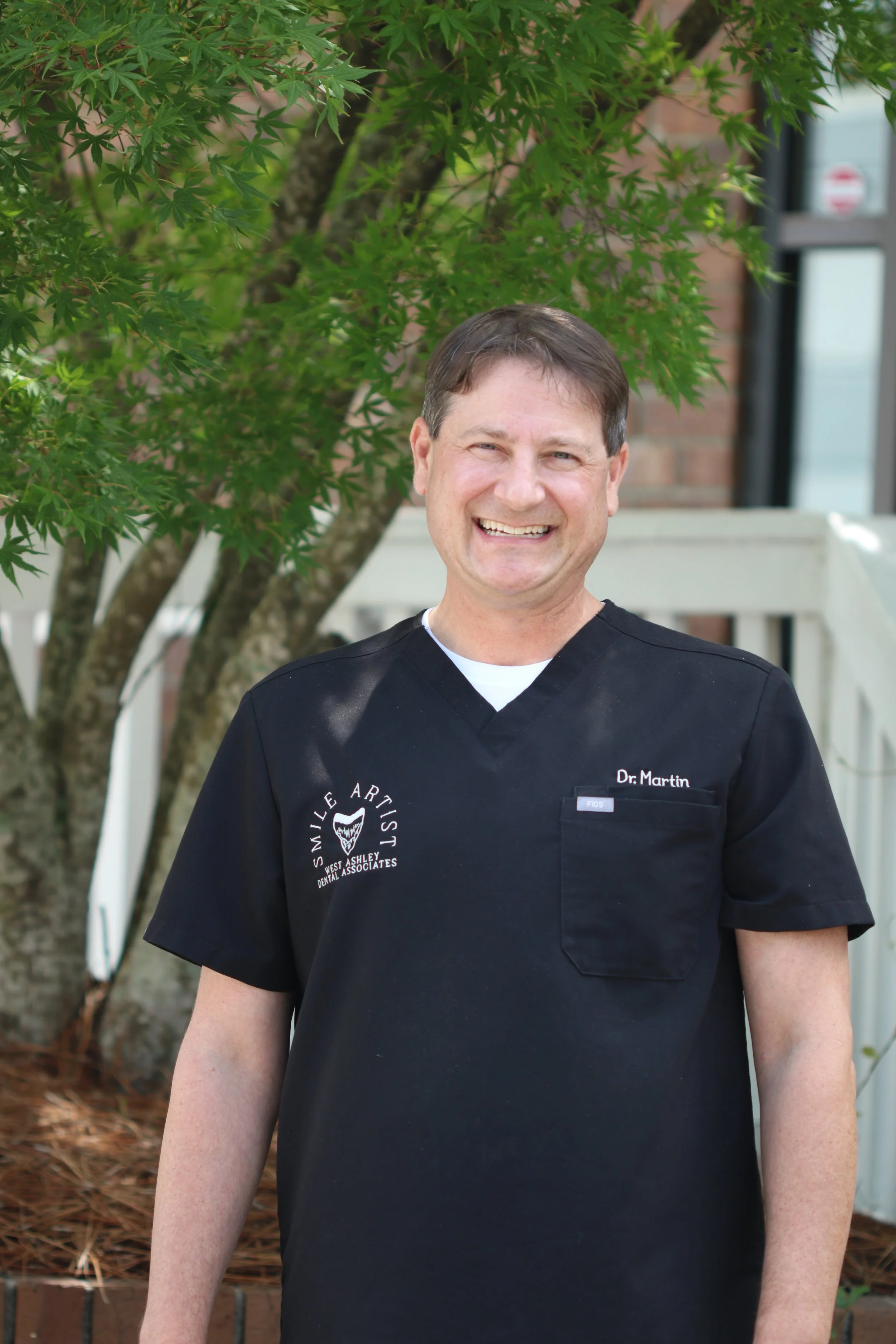 A smiling man wearing black medical scrubs standing outdoors near a tree with green leaves.