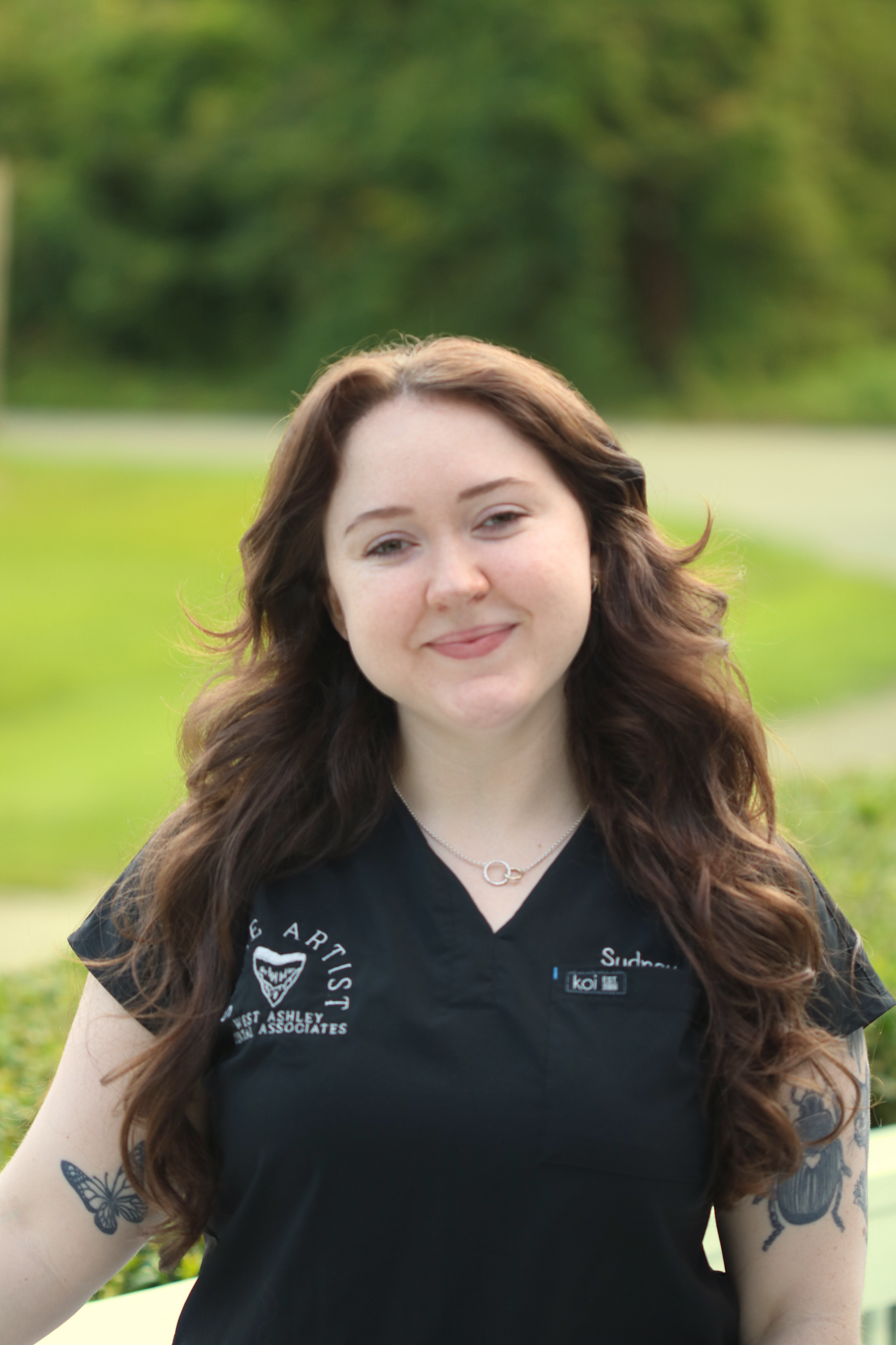 A young woman with long, wavy brown hair and multiple tattoos on her arms, smiling outdoors in front of a blurred green background. She is wearing a black shirt with logos and text on it and a silver necklace.