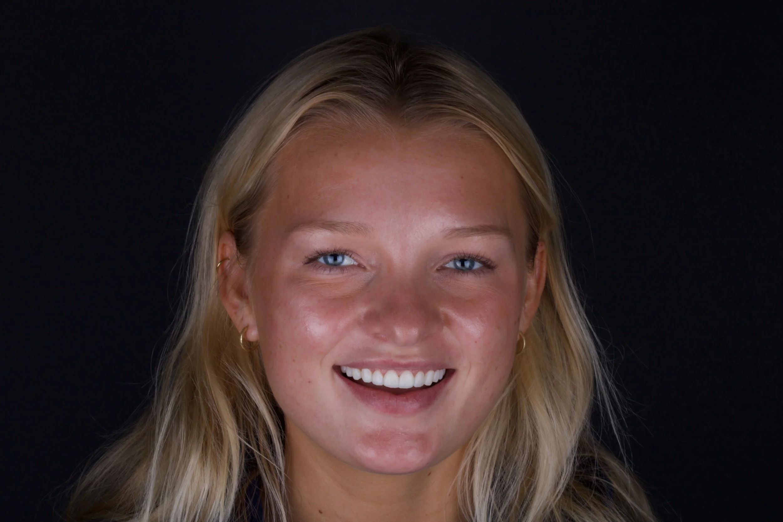 Close-up photo of a smiling woman with blue eyes, blonde hair, and small hoop earrings, against a black background.