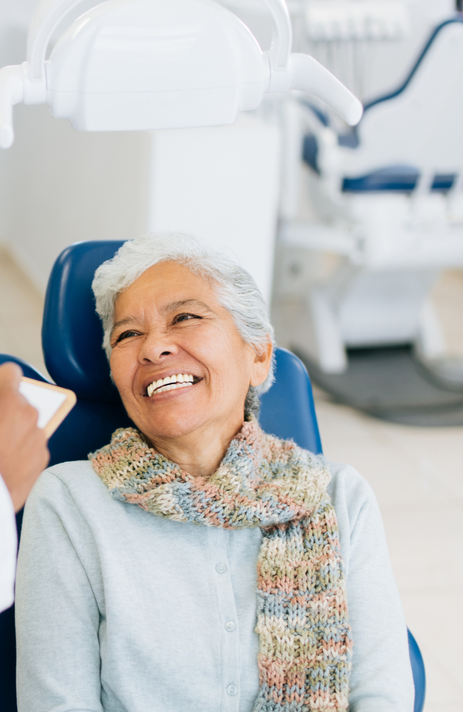 An elderly woman with gray hair wearing a multi color scarf smiling in a dental clinic, sitting in a dental chair, with dental equipment in the background.