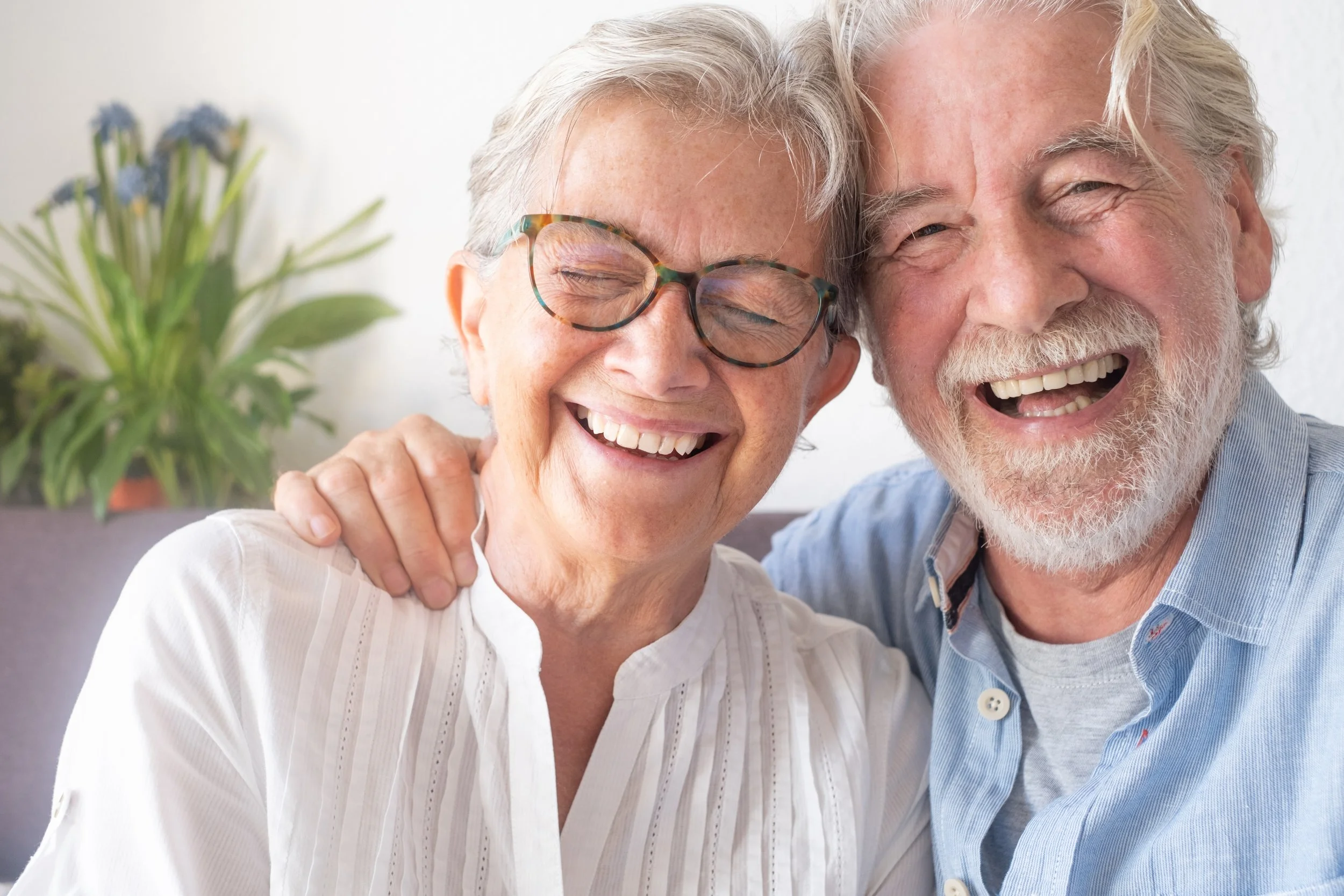An elderly couple smiling and hugging each other, with a fuzzy plant in the background.