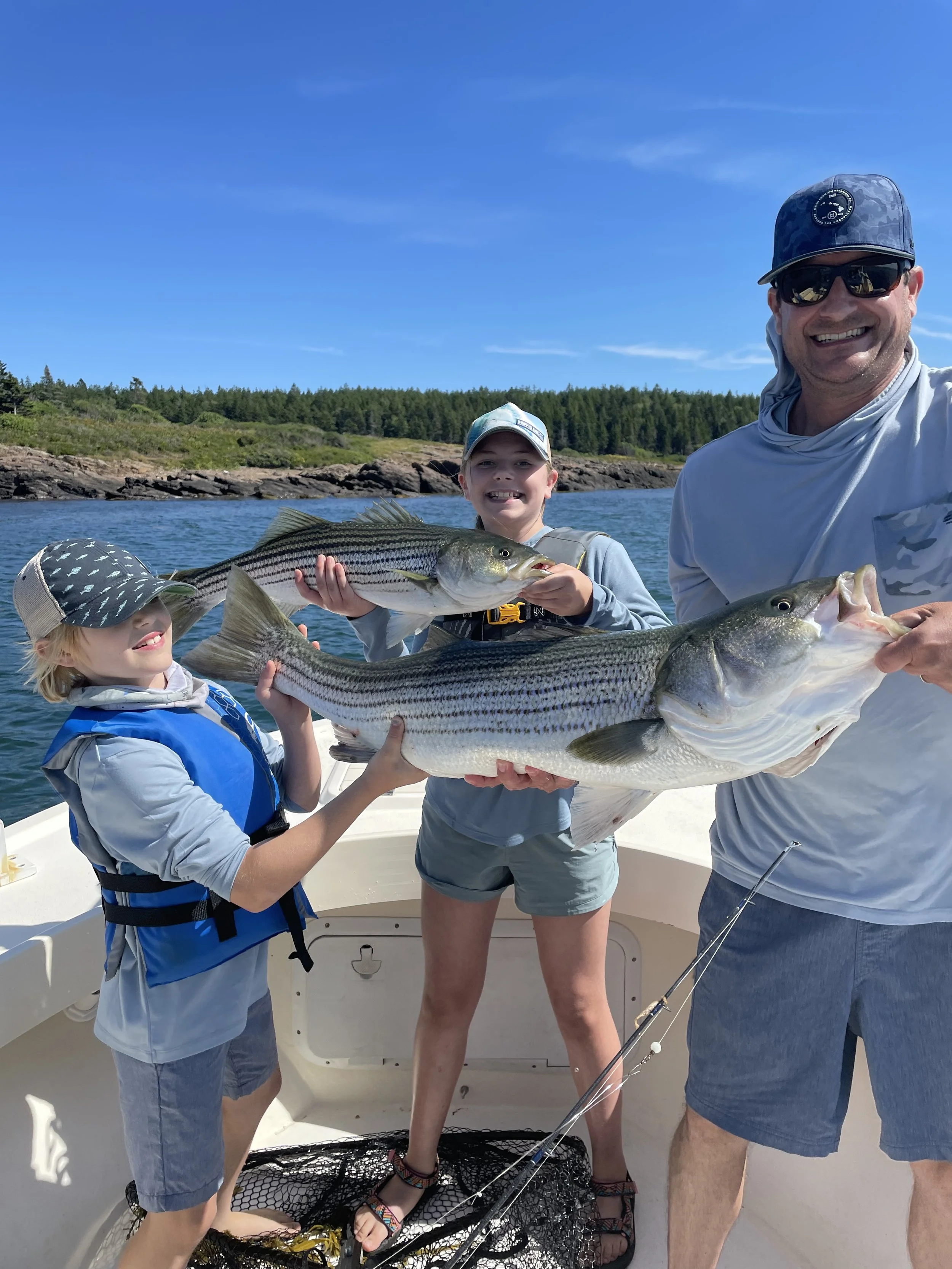 A family on a boat holding large fish they caught, with a scenic background of water, rocks, and green trees under a bright blue sky.