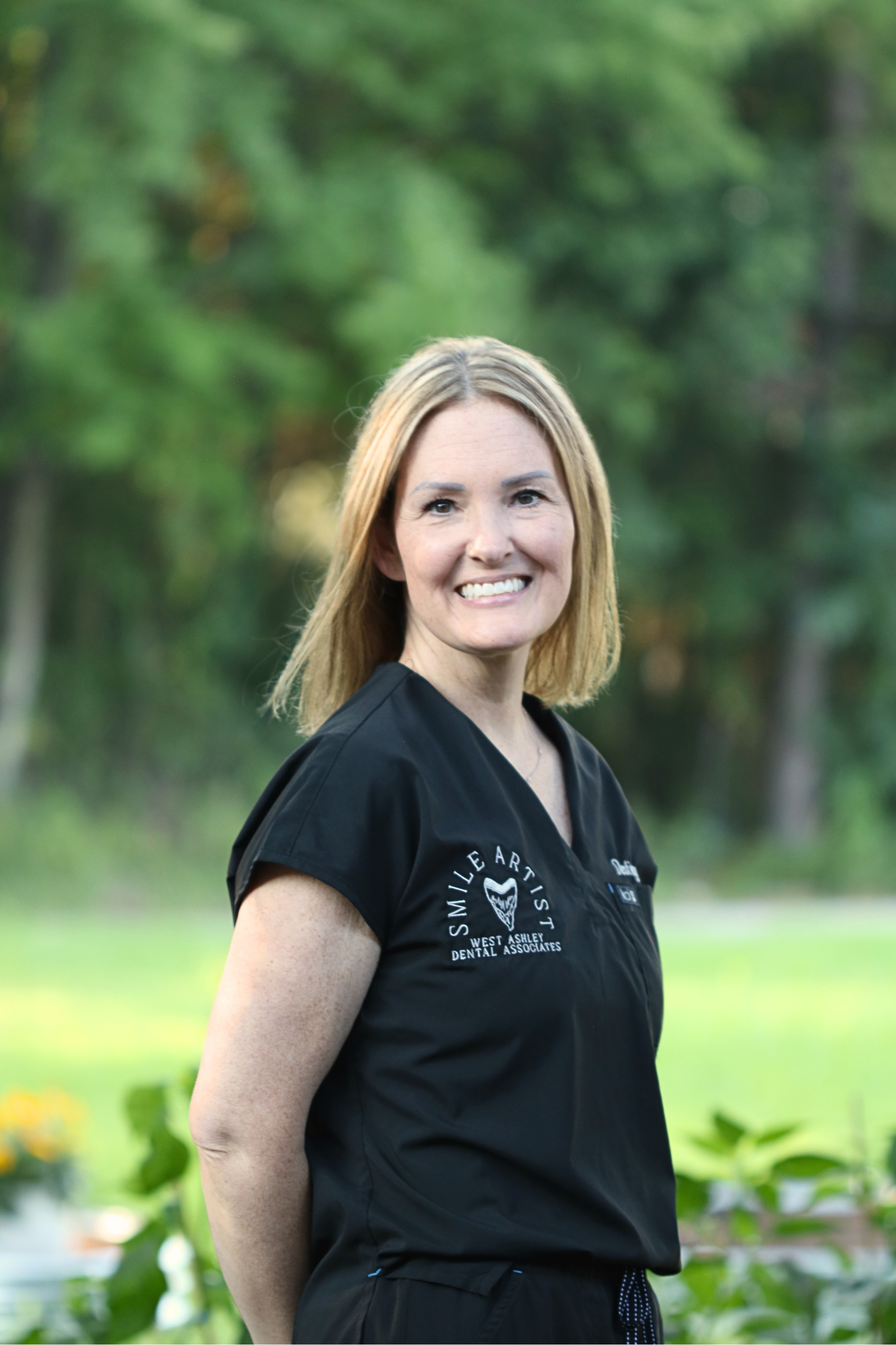 A woman with shoulder-length blonde hair smiling outdoors, wearing a black medical or dental uniform with a logo on the chest, in front of a background of green trees and bushes.