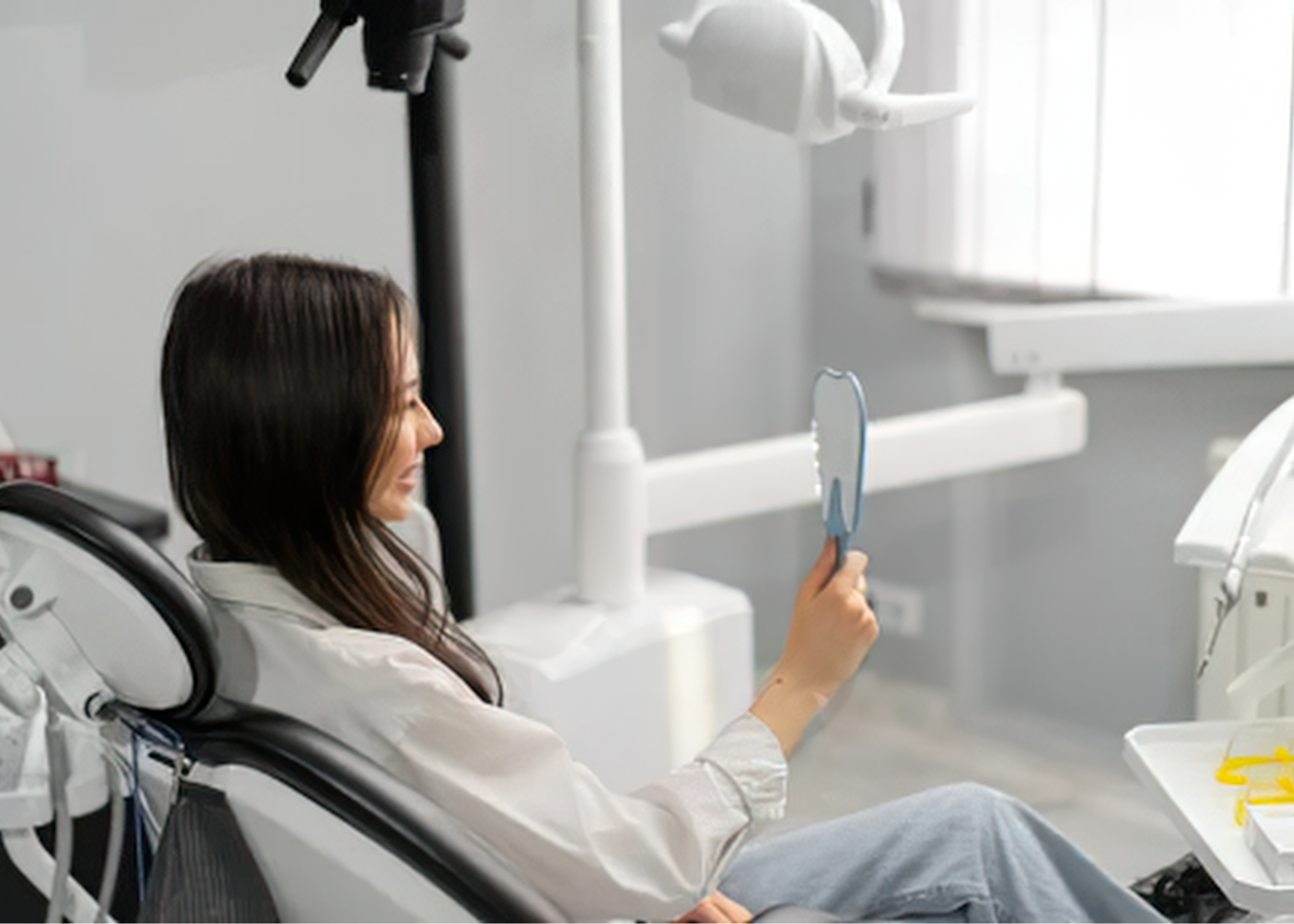 A woman sitting in a hospital bed smiling while holding a dental mirror in her right hand in a dental office or clinic.
